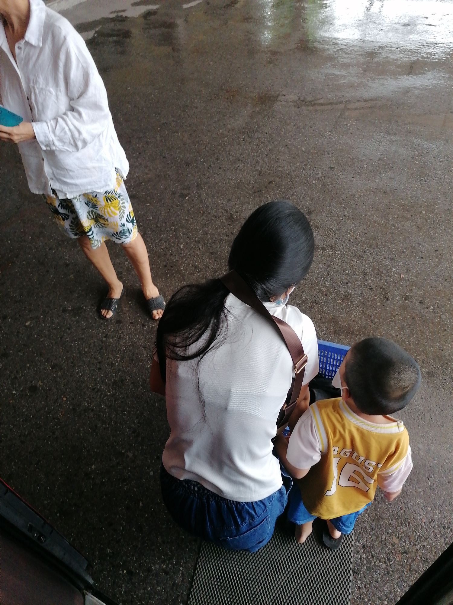 An overhead shot in Chu Văn An Ward, Vietnam, shows three people: a woman, a child, and another person partially visible on the left. The woman is crouching, wearing a white shirt, blue pants, and a brown strap across her shoulder. The child next to her wears a yellow shirt with "AGUS 16" printed on the back and blue pants. Both the woman and child wear face masks. A blue basket sits near the woman. To the left, an individual wearing a white shirt and patterned shorts stands, holding a blue object. The ground is a mix of asphalt and patterned matting. Some areas of the asphalt are wet.
