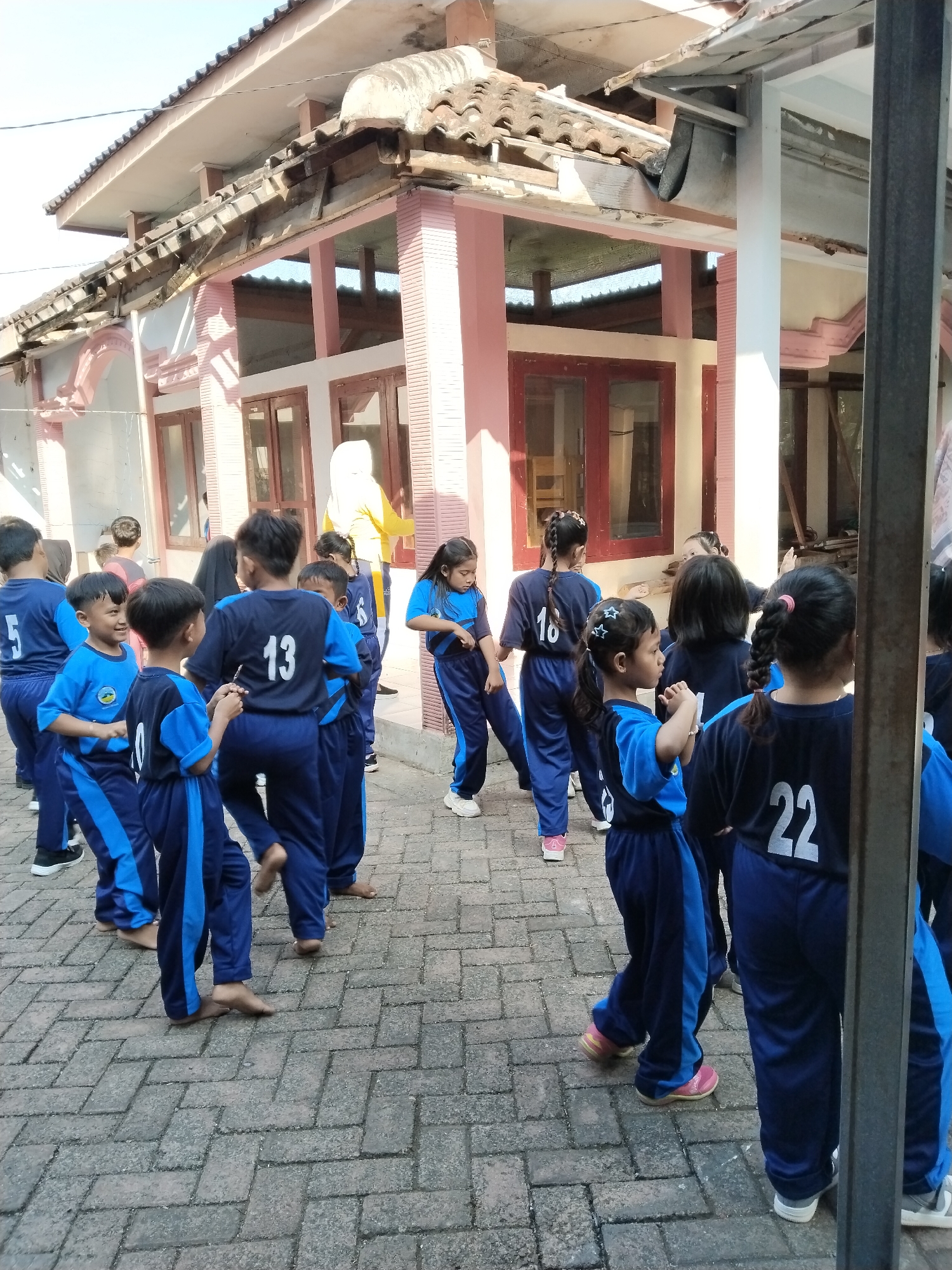 The image depicts a group of children in dark blue and light blue uniforms standing on a brick-paved area outside a building with a pink facade and a tiled roof. Some of the children have numbers on their uniforms, including "13", "16", and "22". One child is wearing pink shoes, while others are barefoot or wearing white shoes. A woman in a yellow hijab and top is visible in the background, interacting with one of the children. The building appears to be a school or similar institution. The lighting suggests it is daytime, with clear sky visible above. This scene is located in Bendoroto, Indonesia, as indicated by the provided context.