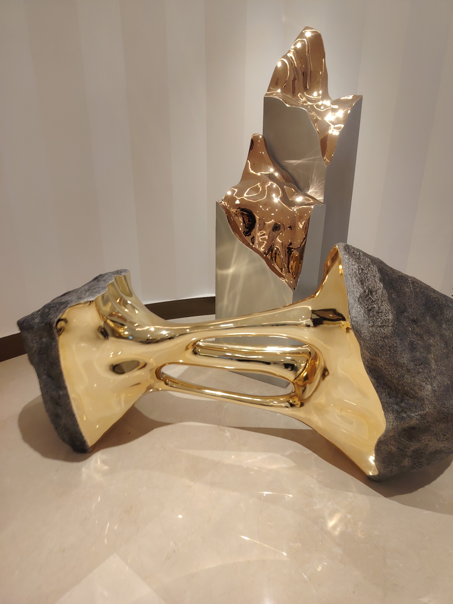 The image depicts a modern art installation featuring two abstract sculptures. In the foreground, a large, rough, grey stone is bisected, with its interior revealing a highly polished, golden, trumpet-like form. This golden element has a smooth, reflective surface with curved, hollowed-out sections, creating a sense of fluid movement. In the background, a taller, grey, rectangular base supports another abstract sculpture made of the same polished golden material. This upper sculpture has irregular, undulating shapes, catching the light and reflecting the surroundings. The sculptures are placed on a light-colored, reflective floor, likely marble or polished concrete, which mirrors the light and forms. The background wall is a neutral, light beige color with subtle vertical lines. There are no people visible in the image. The overall impression is one of contrasting textures and materials, juxtaposing the natural roughness of stone with the artificial smoothness and gleam of polished metal. The location is identified as Konigil, Uzbekistan.
