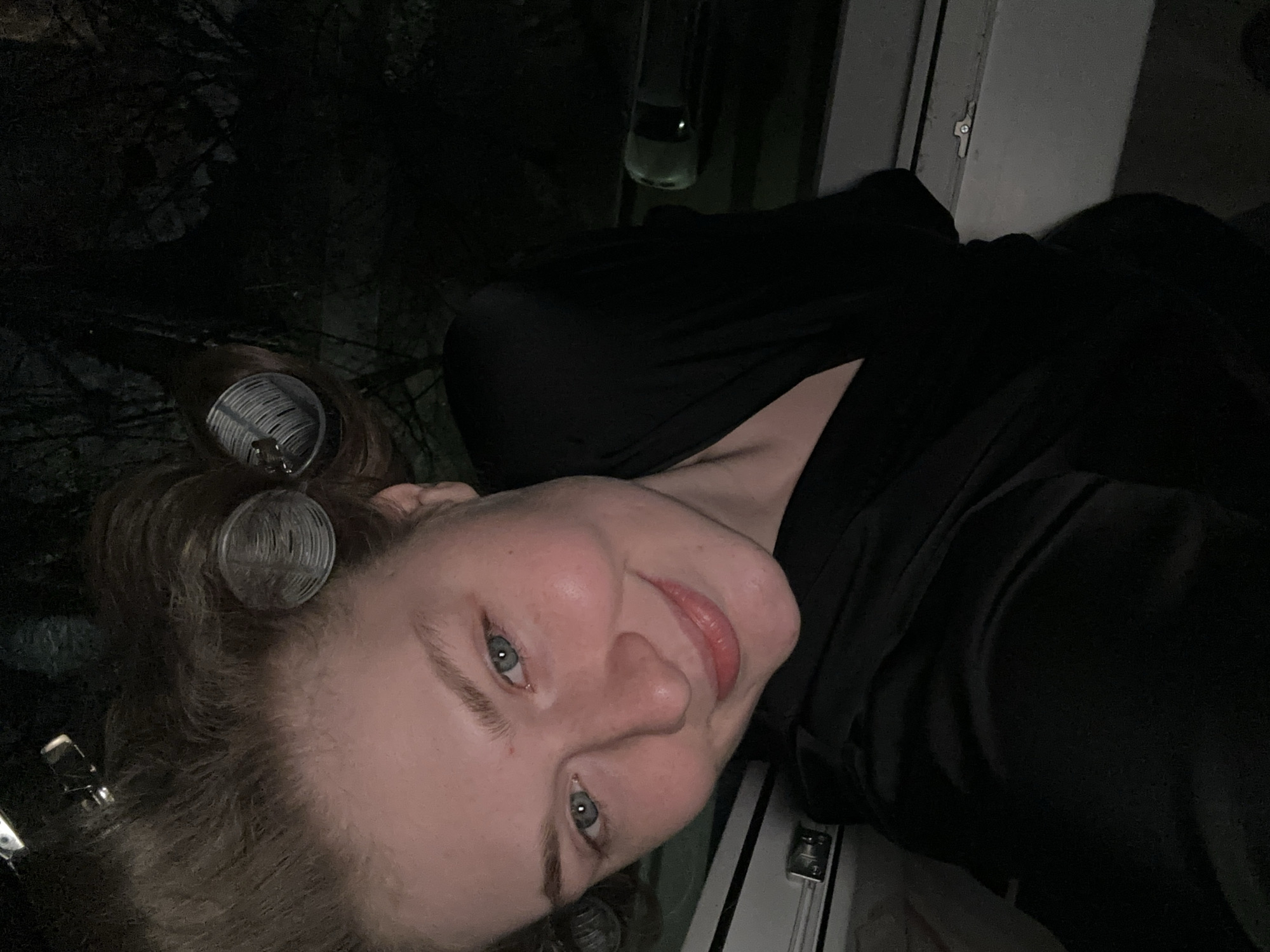 A close-up, slightly angled shot of a fair-skinned person with curlers in their hair, wearing a black garment, and looking at the camera. The person has light blue eyes and a faint smile. To the left, there is what appears to be dark foliage. The background is dark and largely indistinct, with a few undefined light sources. The setting and location within the city of Unknown, Unknown are undetermined.
