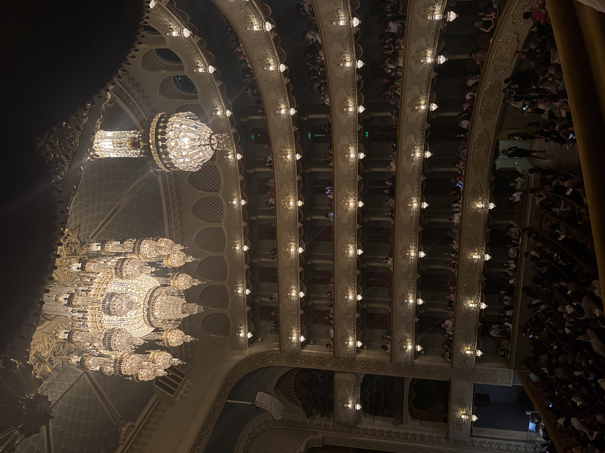 The image depicts the interior of an ornate theatre, likely a performance venue for plays, operas, or concerts. The location is unknown. The focus is directed upwards towards the ceiling, showcasing two large crystal chandeliers. The ceiling is decorated with intricate designs and patterns. The theatre has multiple levels of balconies filled with an audience. The audience members are partially visible, appearing as a mass of people seated in the balconies. Individual audience members are indistinguishable due to the distance and angle of the shot. Lighting fixtures line the edge of each balcony level. The atmosphere is dark, illuminated by the chandeliers and the lighting fixtures. A curtain is partially visible on the right side.