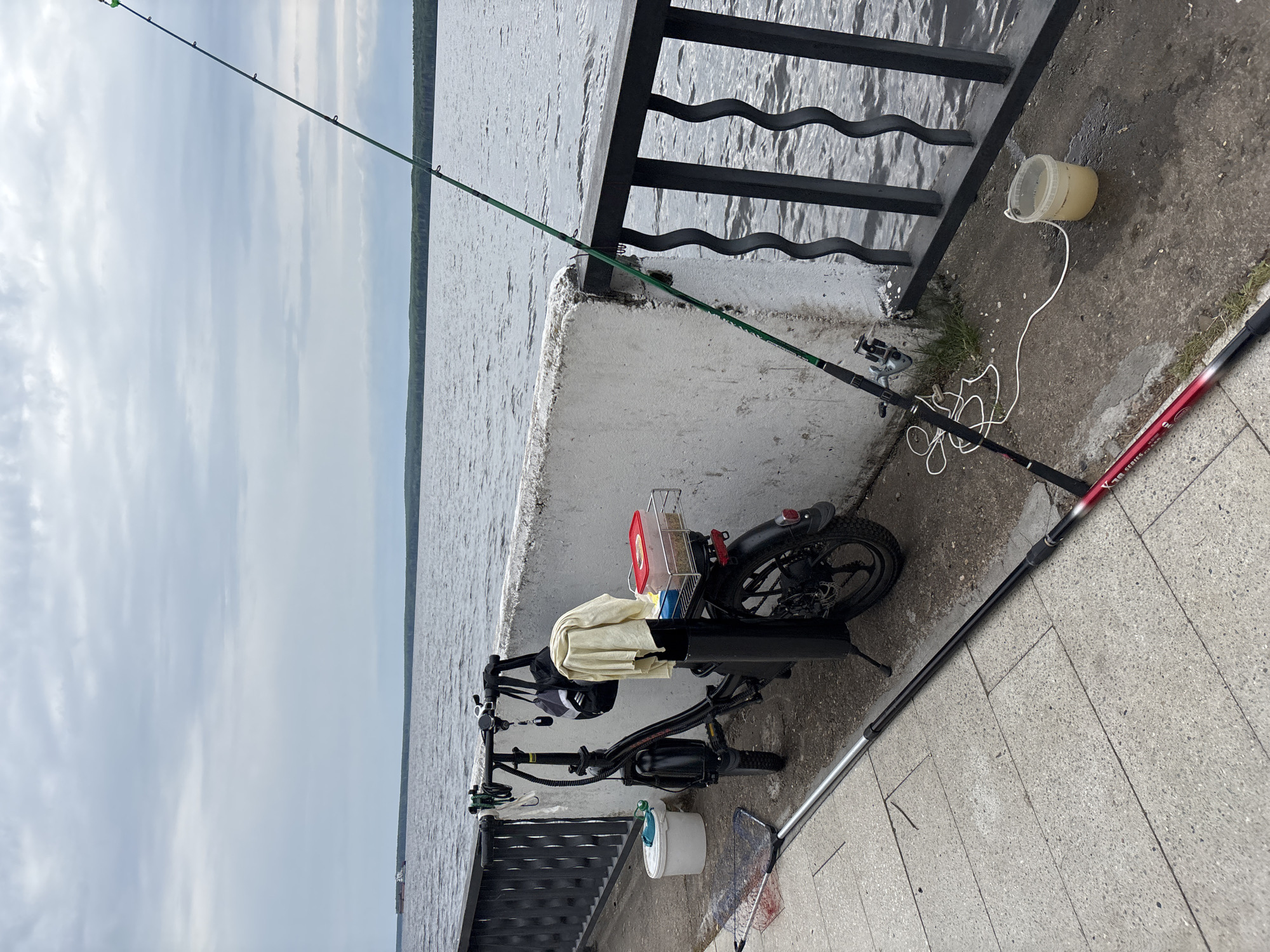 The image, taken in Chaikovsky, Russia, depicts a fishing setup on a concrete bank beside a large body of water. An electric bicycle is parked on the bank, with a cream-colored cloth draped over its handlebars and seat, and a red container and a mesh basket attached to its rear rack.
Several fishing items are present: a green fishing rod with a reel extends from the concrete over the water, actively set for fishing. Another red and black fishing rod, bearing the text "X-TREME SERIES," lies on the concrete. A fishing net with a long handle is also visible on the ground. Two plastic buckets are on the bank: a white one with a lid near the bicycle's front wheel, and a light yellow-beige one, seemingly holding bait or water, positioned closer to the water's edge.
A black metal railing with wavy horizontal bars separates the concrete bank from the water. The expansive water body exhibits gentle ripples under a bright, overcast sky. A distant treeline is visible on the horizon, suggesting a large lake or wide river. The scene portrays a tranquil daytime setting for fishing, with equipment ready for use.