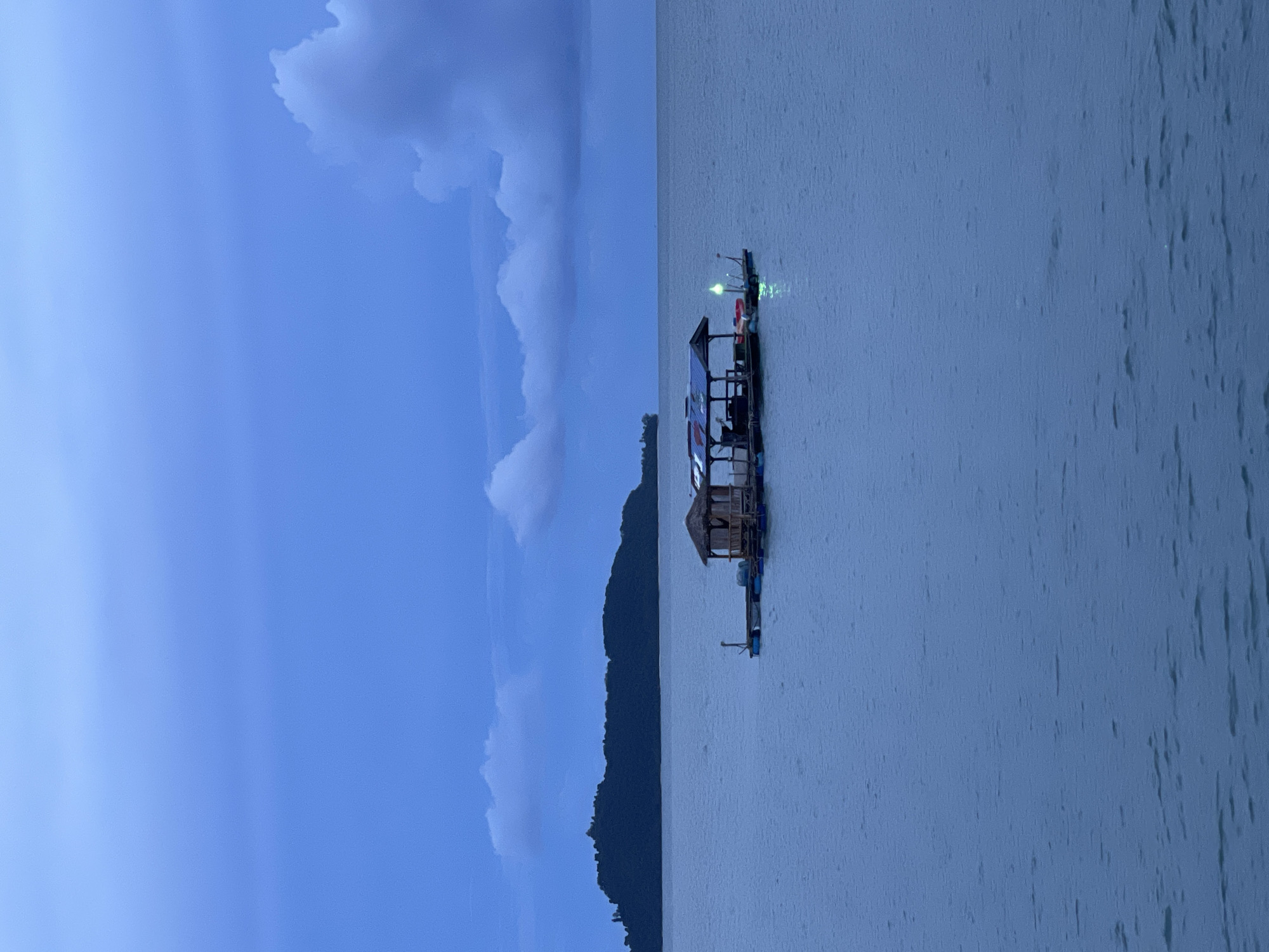 The image depicts a serene scene at dusk in Pa Khlok, Thailand. The primary subjects are a floating structure, possibly a house or raft, on a calm body of water and a silhouetted mountain range in the background. The floating structure is wooden and appears to have a thatched roof on a smaller section, while a larger section has a more modern roof. There are lights visible on the floating structure, suggesting it is inhabited or in use. Small rafts or buoys are also visible near the main structure.
The time of day is clearly dusk, indicated by the deep blue hues of the sky and the soft, diffused lighting. The weather appears to be calm, with the water showing minimal ripples. The sky is partially filled with large, soft clouds, adding to the tranquil atmosphere. The mountain range's dark silhouette against the twilight sky provides a sense of depth and natural beauty to the scene.
The overall mood of the image is peaceful and contemplative. There is no visible text in the image. The scene captures a quiet moment, likely at the end of a day, with the lights on the floating structure offering a hint of human presence in the otherwise natural landscape. The location context of Pa Khlok, Thailand, suggests a tropical or subtropical setting.