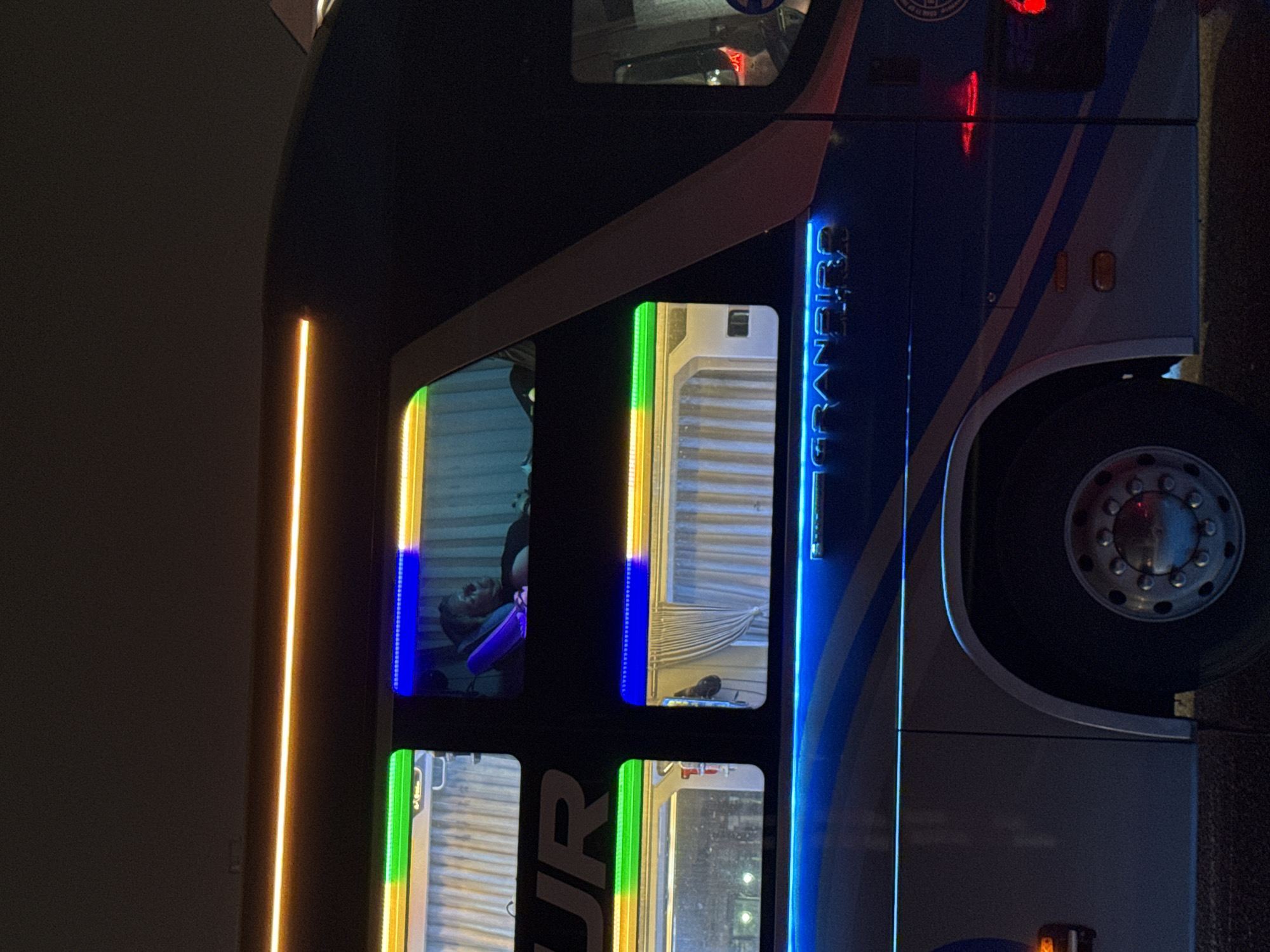 The image shows the side of a passenger bus at night. The bus has large windows framed with colored lights, and a lit sign that reads "GRANDBIRD TOUR." The color scheme transitions from blue to yellow to green in the window frames. A large tire is visible. The bus is near a wall with a vertical orange light fixture. It's not possible to confirm the image was taken in Đà Nẵng, Vietnam, with the given context.