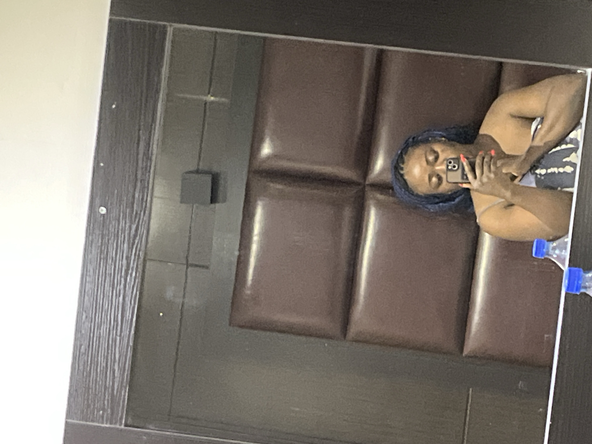 A mirror reflection shows a woman taking a selfie with a smartphone. She has blue braided hair, dark skin, and red nail polish, and is wearing a light-colored top. Behind her in the reflection is a brown, padded headboard. A black cube object is attached to the wall to the left of the headboard. To the right, two water bottles are visible. The mirror has a dark, wood-like frame. Based on the interior setting, and the mention of Abuja, Nigeria, it may be a room in a residential or commercial building, but the specific location is not clear from the image.