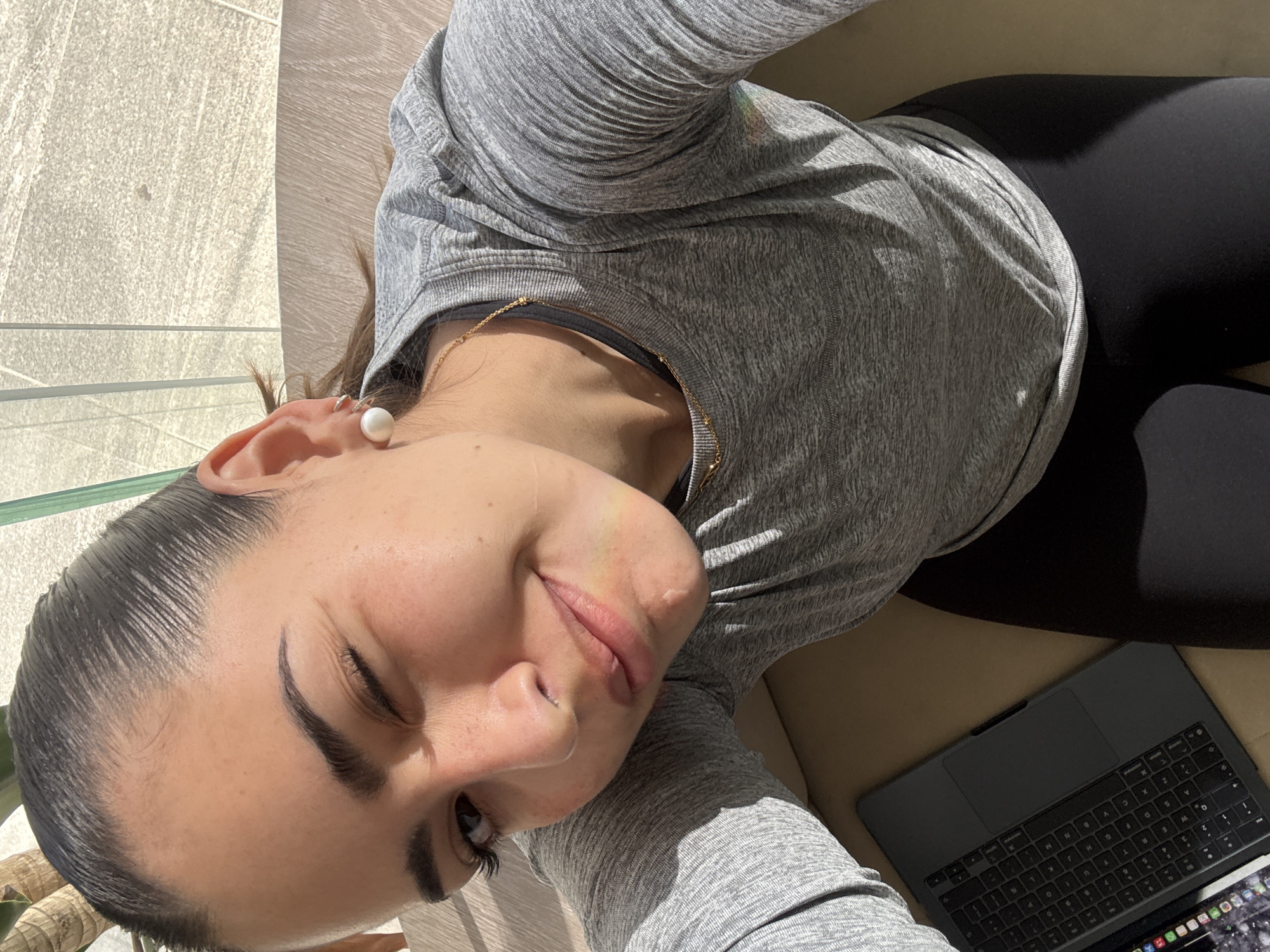 This image captures a close-up, high-angle view of a young person, likely female, lounging in a sunny setting in Puebla, Mexico. The individual is dressed in a grey, long-sleeved athletic top and black leggings. A notable detail is a large, spherical white earring worn on the visible ear, complemented by a delicate gold chain necklace. Her dark hair is neatly pulled back, and she gazes towards the camera with a relaxed expression, one eye partially closed in a soft smile, possibly due to the bright light or a contented mood.
The scene is bathed in strong, natural sunlight, indicated by pronounced shadows cast across the clothing and surfaces. The background reveals a textured, light-colored wall or building facade and a transparent glass railing, suggesting an outdoor balcony, patio, or a room with large windows. Beside the person, a dark grey laptop, resembling a MacBook with a protective case, rests on a light brown or tan upholstered surface, indicating a moment of work or leisure. The bright conditions suggest it is daytime and sunny.
The visible text from OCR primarily consists of scattered alphanumeric characters, "D", "A", "W", "E", "R", "2", "0", "S", "X", "6", "7", "A", "G", "H", "K", "L", "M", "P", "N", which are likely detected from the laptop's keyboard.