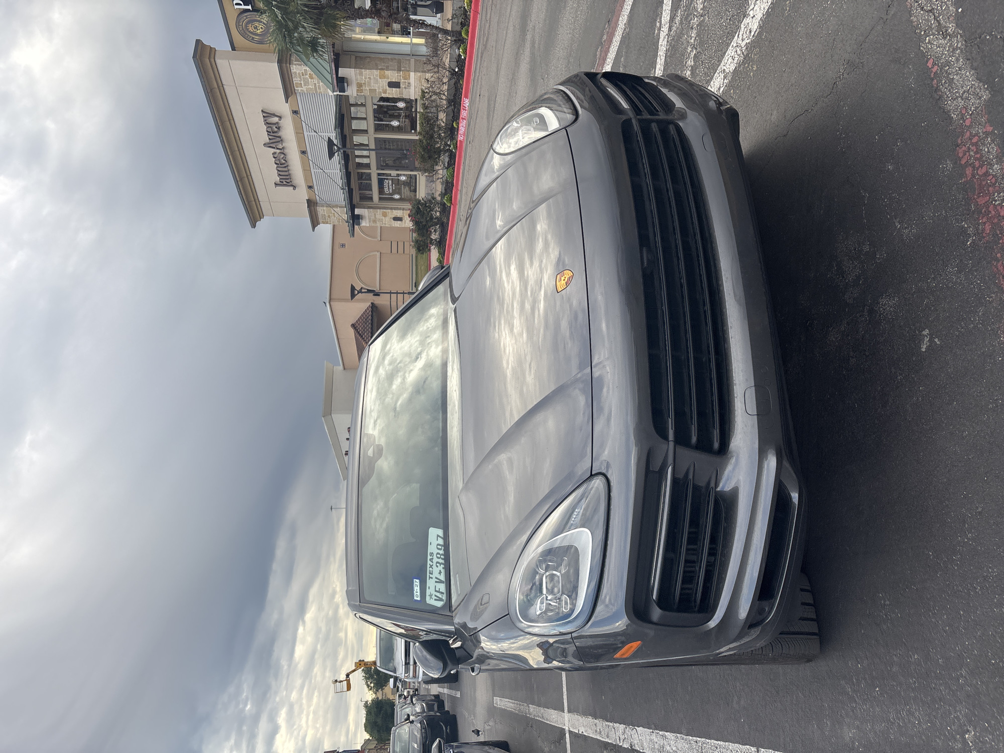 The image shows a dark gray Porsche Cayenne parked in a parking lot in front of the James Avery store in San Marcos, Texas. The car has a Texas license plate, and the sky is cloudy. Other vehicles are parked nearby.