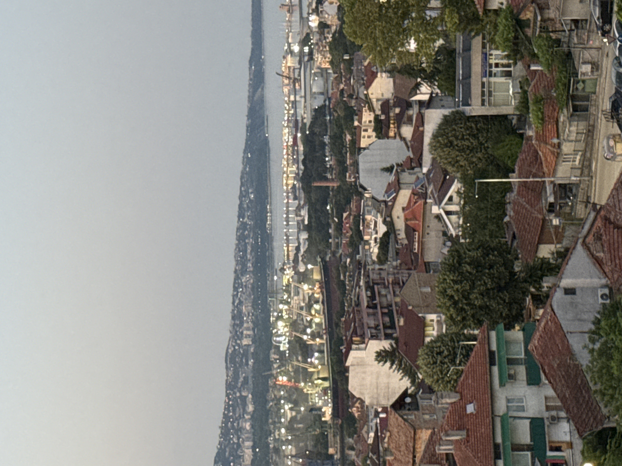 This is a vertically oriented, high-angle photograph looking down on a dense urban area situated alongside a body of water. The scene appears to be a city or town with a mix of residential and industrial buildings. The time of day is likely dusk or early evening, as many lights are illuminated within the buildings, creating a warm glow. The sky is overcast and a pale gray, suggesting a cloudy day or the approach of night.
The architecture visible includes numerous red-tiled roofs, typical of many European cities. Some buildings are multi-story, with visible windows and balconies. Green trees are interspersed throughout the urban landscape, adding a touch of nature to the built environment. In the distance, along the waterfront, industrial cranes and structures are visible, suggesting port activity. The water itself is a dark, calm expanse stretching into the horizon.
The overall mood of the image is one of a bustling, lived-in city at the end of the day. There are no discernible people or specific events taking place, but the lit windows suggest ongoing activity within the homes and businesses. The location is not specified, but the architectural style and the presence of a significant body of water hint at a coastal or riverine city. No text is visible in the image.