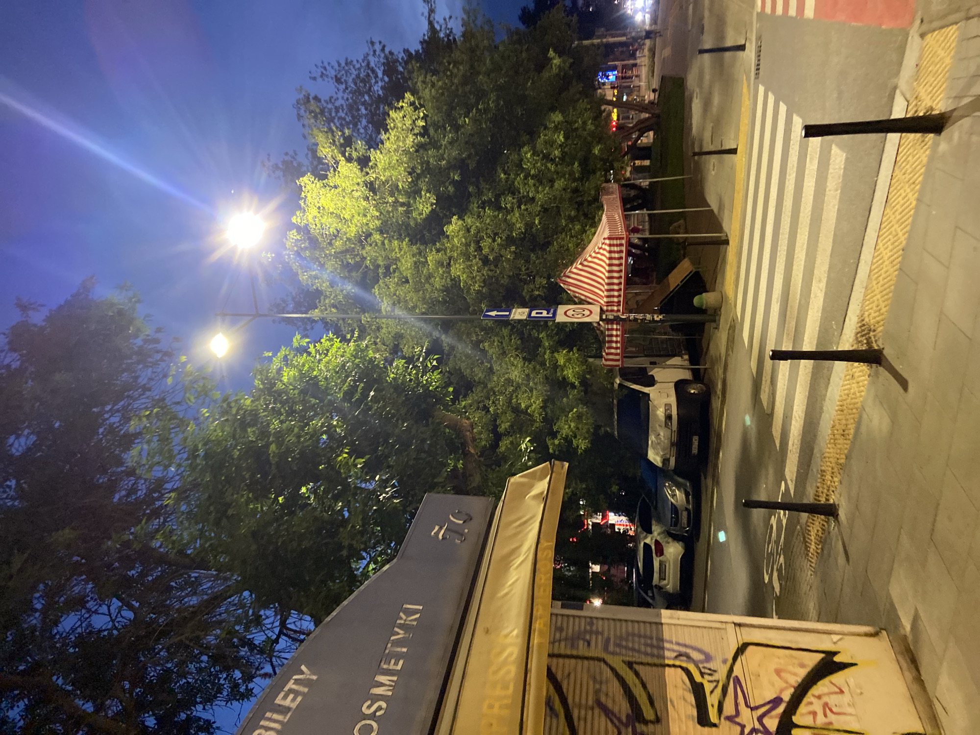 This image depicts an outdoor scene at dusk or night in Warsaw, Poland. In the foreground, a pedestrian crosswalk with tactile paving is visible. To the left, a building facade with graffiti and a yellow awning is present. Text on the awning reads "PRESSA" and the building signage above states "BILETY COSMETYKI". Further into the scene, several vehicles are parked, including a white van and cars. A red and white striped tent with a play slide is set up near a street lamp and traffic signs, one indicating a speed limit of 30. Large trees with illuminated foliage dominate the background. The sky is a deep blue, suggesting the twilight hours. There are no people clearly visible interacting in the scene.