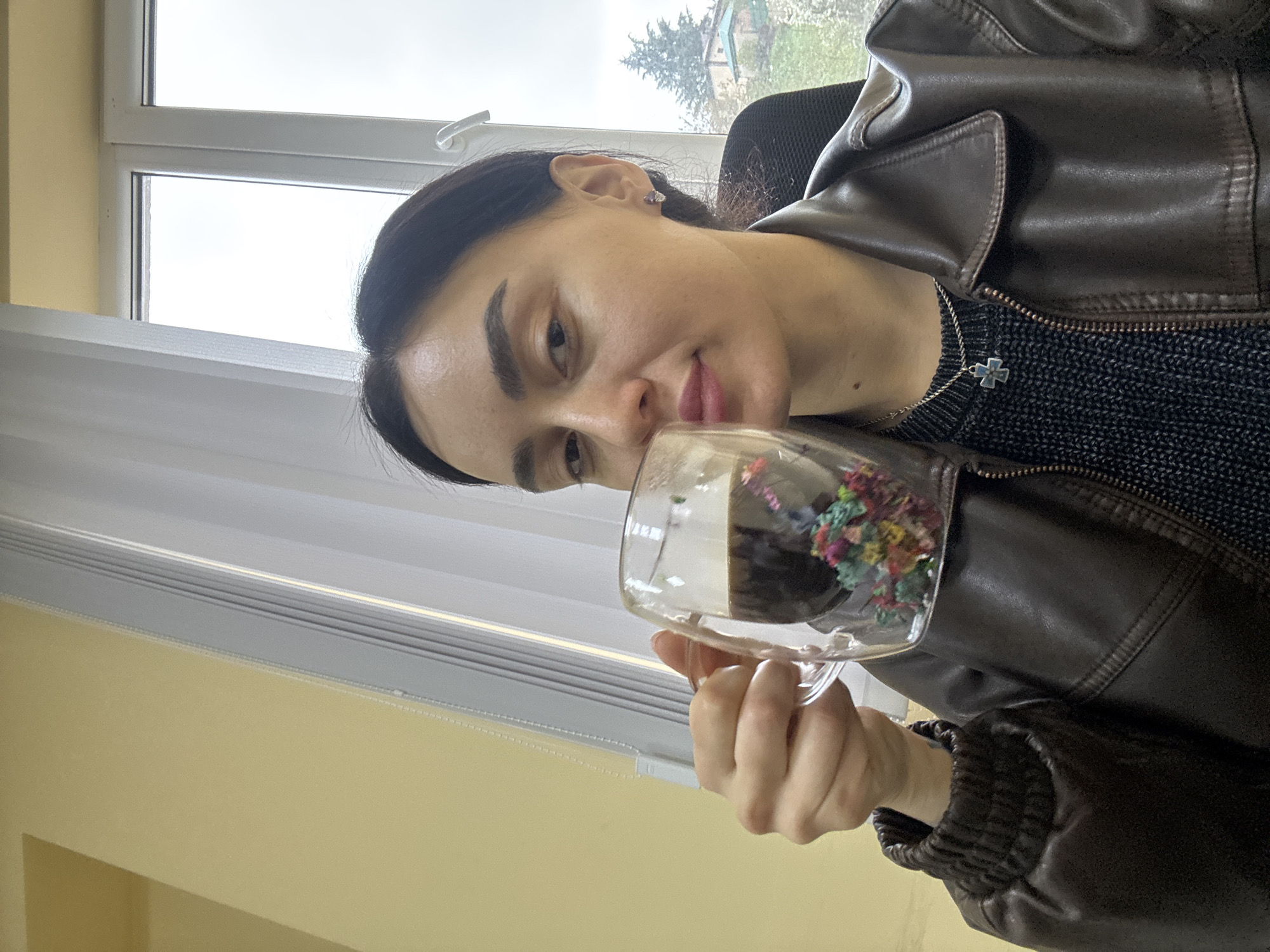 A young woman with dark hair and fair skin is holding a clear glass mug filled with a dark liquid and colorful flower-like decorations. She is wearing a dark, textured top underneath a brown leather jacket with a zipper. A silver necklace with a cross pendant is visible around her neck. She is indoors, possibly near a window with white blinds and a white frame, through which a tree and a distant building can be seen. The wall behind her is a light yellow color.
The location cannot be determined to be specifically Tbilisi, Georgia, without more information. The image is a selfie-style shot taken at a close angle.