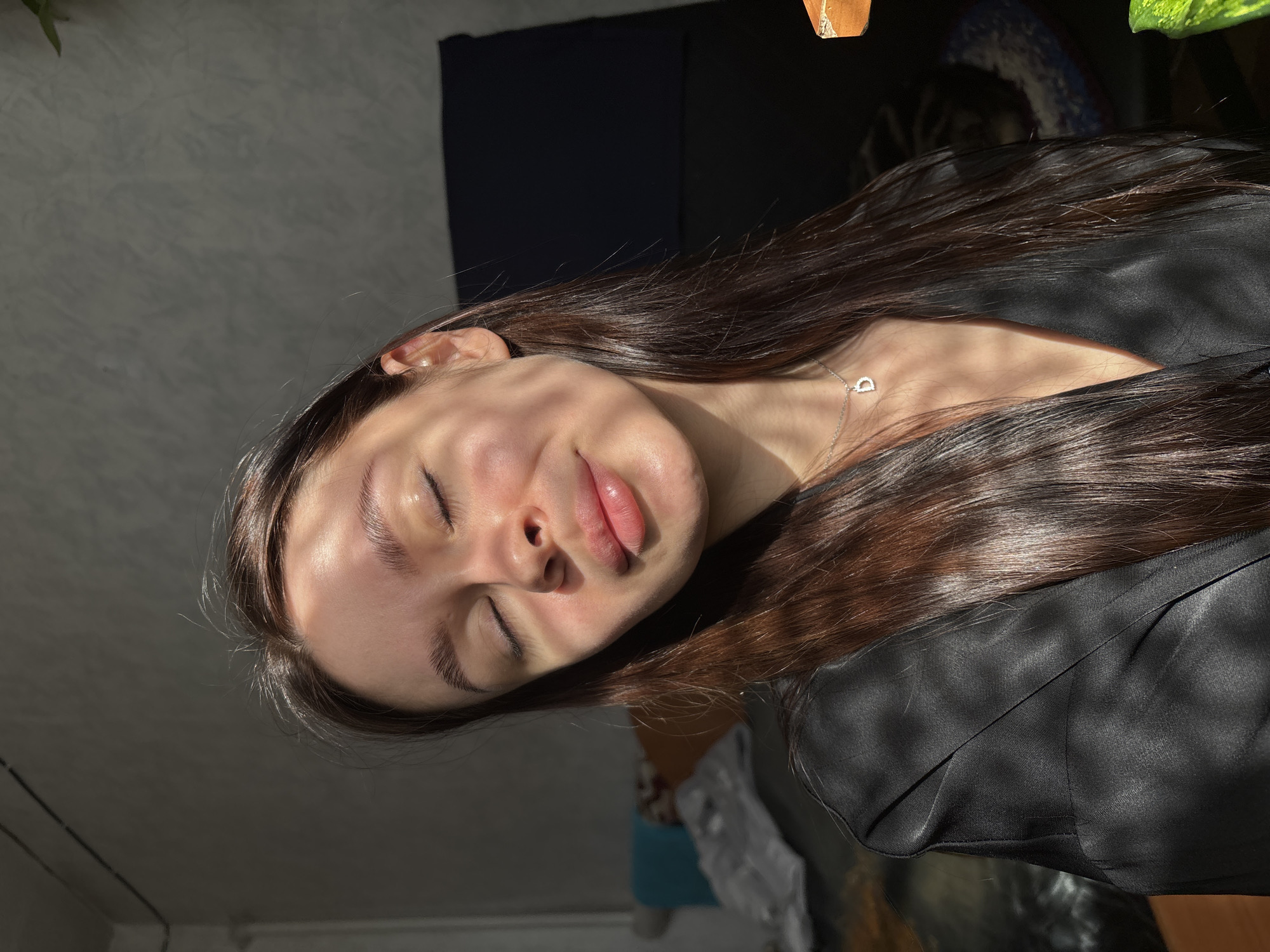 A portrait of a young woman with long brown hair wearing a black shirt, and a silver necklace with a heart-shaped pendant. She has her eyes closed and is facing upwards. Sunlight casts shadows across her face. The background includes a dark wall, a plant, and a glimpse of a room with a wall and possibly a piece of furniture. No information regarding the location in the city of Dubrova, Russia is available.
