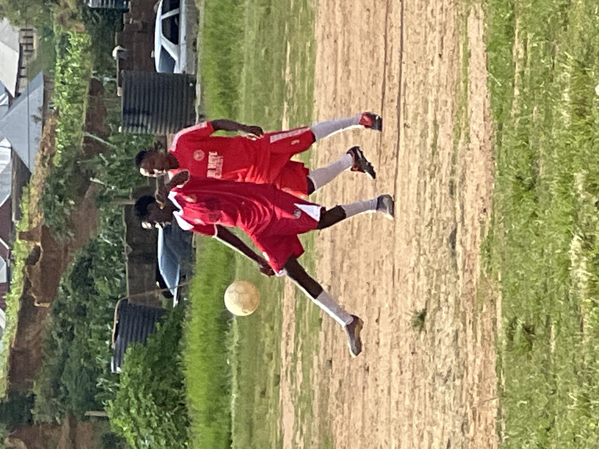 The image depicts an outdoor soccer game. Two young men, dressed in red soccer uniforms with white socks, are visible on a dirt and grass field. The player on the left is in mid-action, kicking a light-colored soccer ball with his right foot. His left leg is extended backward for balance. The player on the right is positioned behind and to the right of the first player, facing the same direction and appearing to observe the play. In the background, there are structures resembling houses with corrugated metal roofs, greenery, and large black water tanks. The overall setting suggests a semi-urban or rural environment, consistent with parts of the city of Jos, Nigeria. The lighting indicates it is daytime.