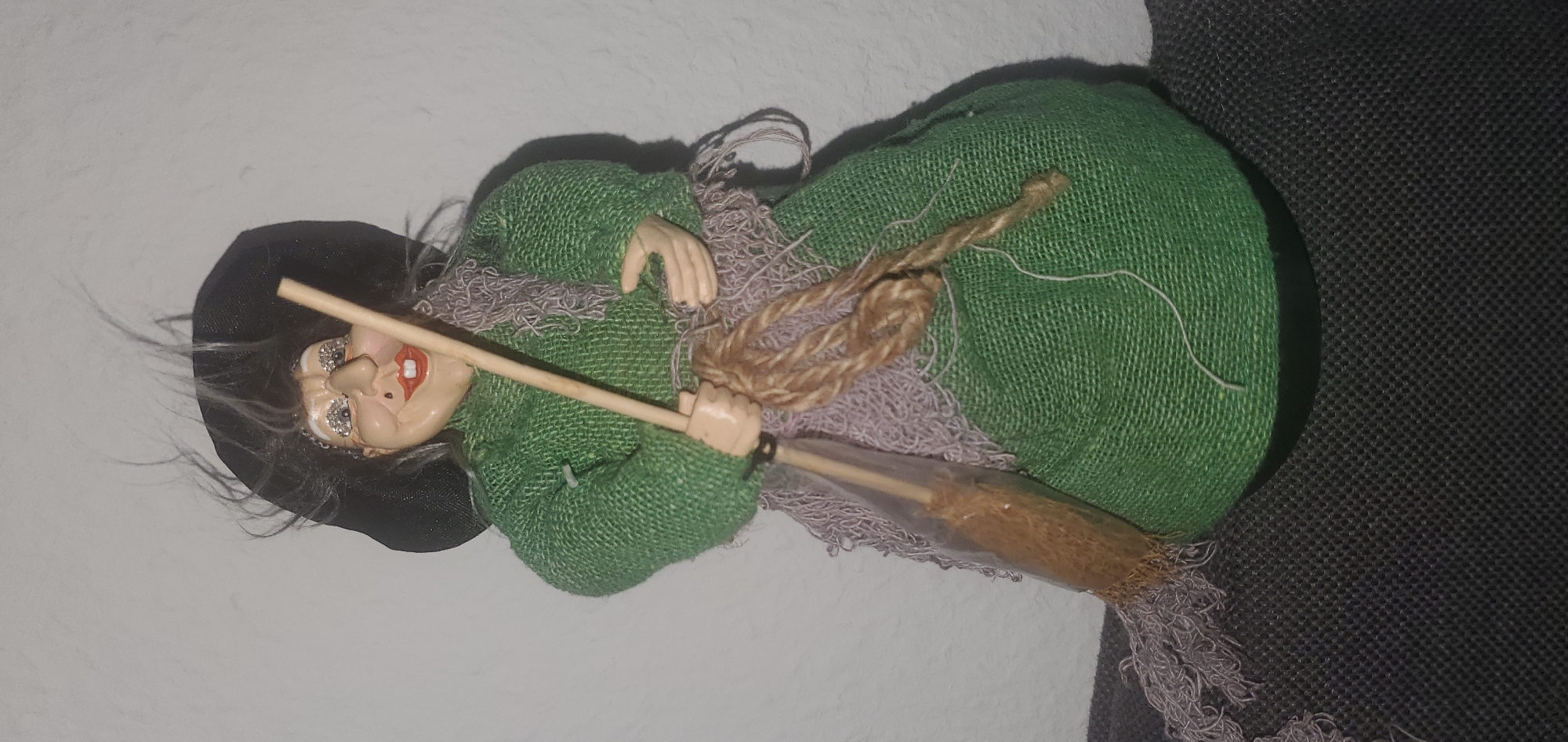 An articulated doll, designed to resemble a witch, stands upright against a textured off-white wall. The doll is wearing a textured green dress and a black hat. It has grey hair that appears to be wispy. The doll's face is painted with exaggerated features, including wide eyes with sparkly accents, a prominent nose, and red lips. In its hands, the doll is holding a broomstick with a light brown handle and straw-like bristles enclosed in clear plastic. A textured, light purple scarf is draped around the doll's shoulders, and a thick, twisted rope is tied around the broomstick. The doll is positioned on a dark grey, textured surface. The image does not provide any information or context related to the city of Sasbach, Germany.