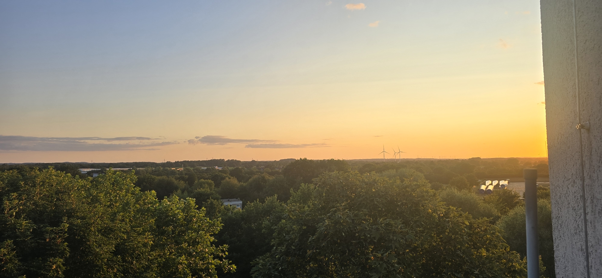 The image shows a sunset over a wooded area with some industrial elements visible in the distance. The sky transitions from a pale blue at the top to a vibrant orange and yellow near the horizon. Several wind turbines are visible in the middle distance, with more silhouetted further away. In the foreground, lush green trees fill the lower portion of the frame. To the right, the textured wall of a building is visible, partially obscuring the view of the landscape. A pipe or conduit runs vertically along the building wall. There are no people or animals visible in the image. This scene is likely captured from a vantage point within or overlooking the city of Kiel, Germany, given the presence of wind turbines which are common in the region.