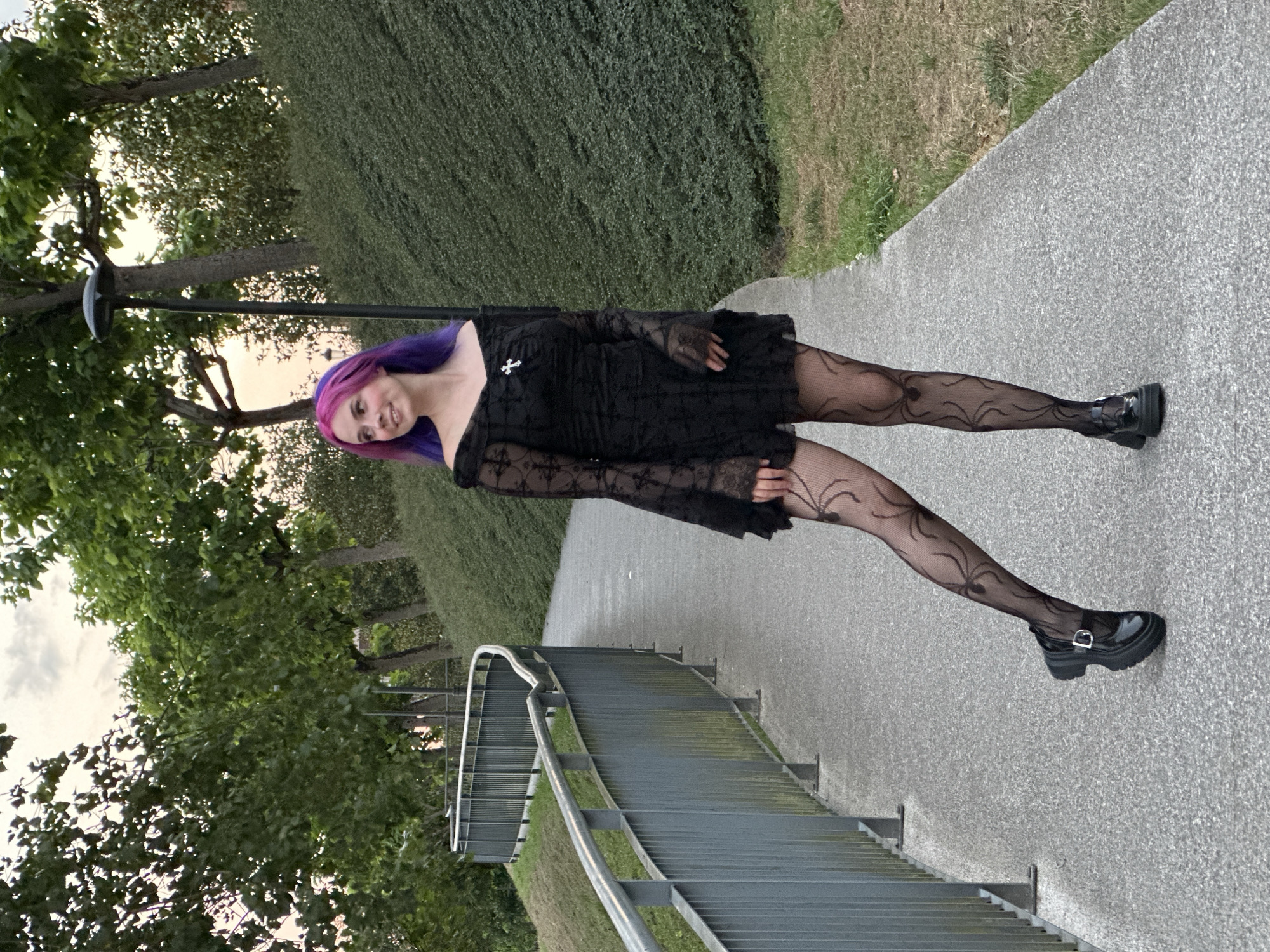 A young woman with purple and pink hair stands on a paved path outdoors. She is wearing a black off-the-shoulder dress with lace detailing, black fishnet stockings with a spider web pattern, and black platform shoes with a buckle. She is looking to her left with a smile. Behind her, a metal railing runs along the edge of a grassy incline. Trees and a hedge are visible in the background. The lighting suggests it is late afternoon or early evening.