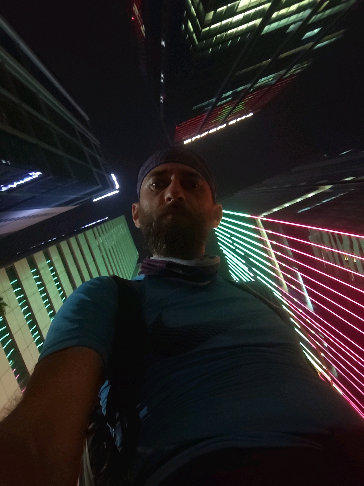 A low-angle, wide shot captures a man wearing a blue t-shirt and a dark blue baseball cap, looking upwards. The man has a beard and a bandana around his neck. He is positioned in the foreground, with the background dominated by the brightly lit facades of modern skyscrapers at night. The buildings are illuminated with various colored lights, including neon pink and green, creating a dynamic and futuristic urban landscape. The perspective suggests the photo was taken from a street-level or slightly elevated position, looking up towards the towering structures. The dark sky contrasts with the vibrant illumination of the city. The scene is indicative of a modern urban environment at night, likely in a city known for its impressive architecture and nightlife. This scene is consistent with an outdoor setting in a major metropolitan area like Ho Chi Minh City, Vietnam, known for its vibrant nightlife and modern architecture.