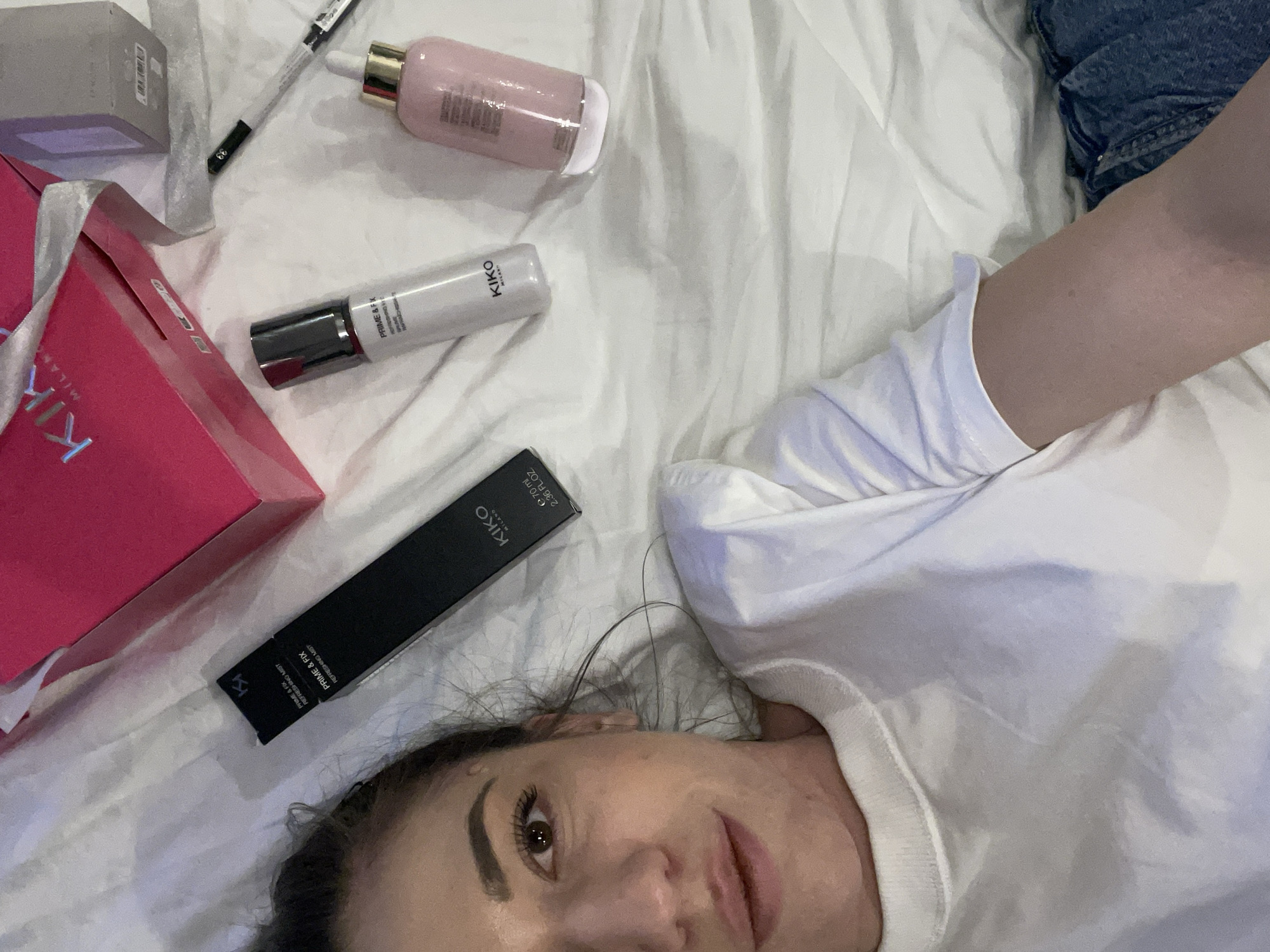 A female individual is depicted lying on a white, wrinkled fabric surface, likely a bed or similar soft furnishing, posing for a photograph. She has fair skin, dark brown eyes, and dark hair pulled back, with a faint smile on her lips. She is wearing a white crew-neck t-shirt and blue denim jeans; her left arm is visible extending downwards. Arranged around her on the fabric are several cosmetic products and packaging. These include a black rectangular box labeled "KIKO MILANO PRIME & FIX REFRESHING MIST E 70 ml 2.36 FL.OZ.", a white pump bottle of the same "KIKO MILANO PRIME & FIX REFRESHING MIST," and a light pink bottle with a gold dropper top. A pink shopping bag bearing the "KIKO MILANO" logo with silver ribbon handles, a small gray rectangular box (also KIKO MILANO branded), and a black eyeliner pencil marked "33" are also present. The lighting is even, suggesting an indoor setting. The precise location within Sofia, Bulgaria, cannot be identified solely from the visual elements within the photograph.