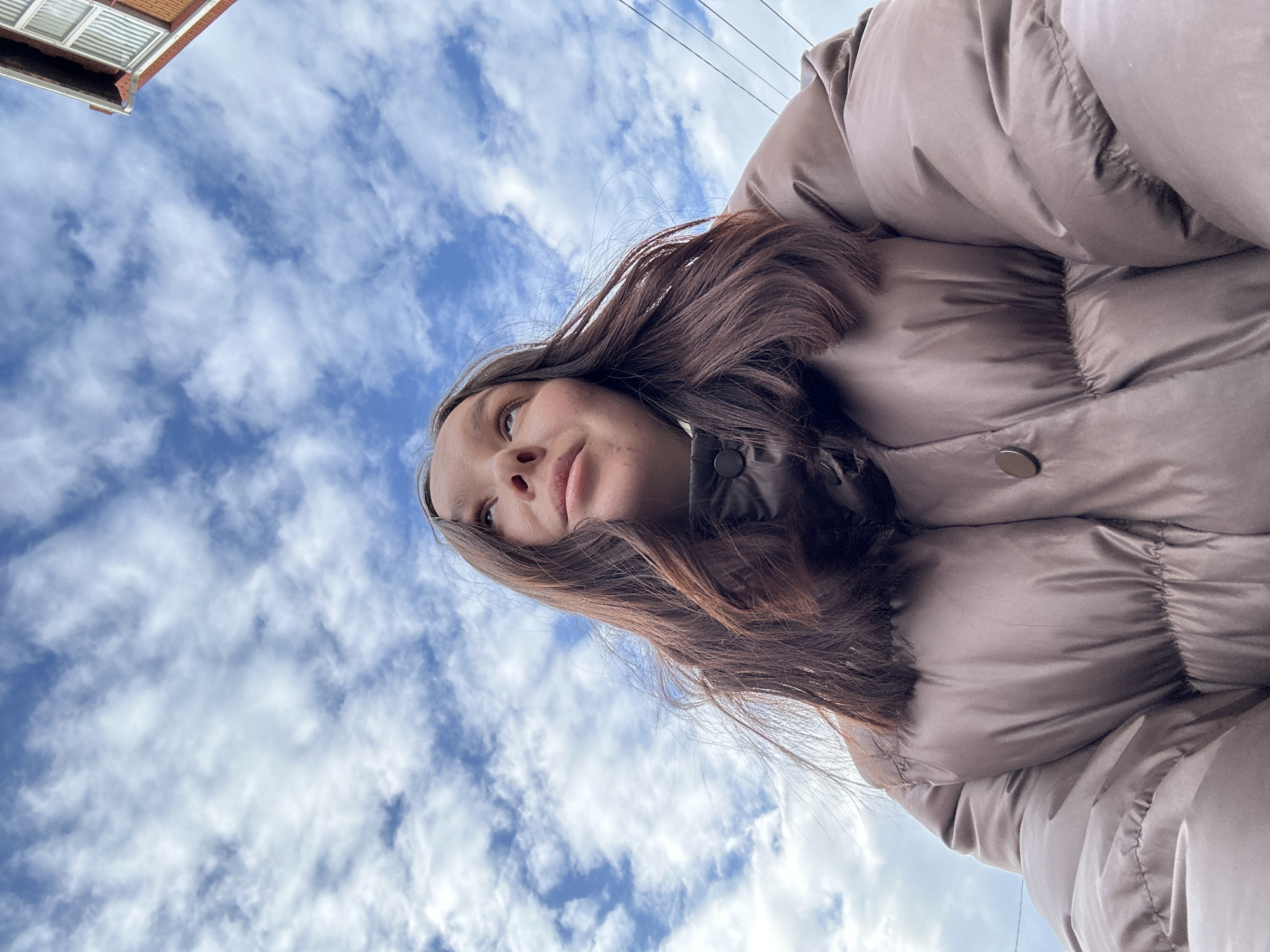 An upward-angled outdoor selfie depicts a young female from the chest up, her gaze directed skyward and slightly right. She wears a light brown or taupe puffy winter jacket. Her long, dark brown hair frames her face. The background features a blue sky with scattered white clouds. A segment of a reddish-brown brick building with white window frames and dark utility wires is visible in the upper right quadrant. This scene is located in Chaikovsky, Russia.