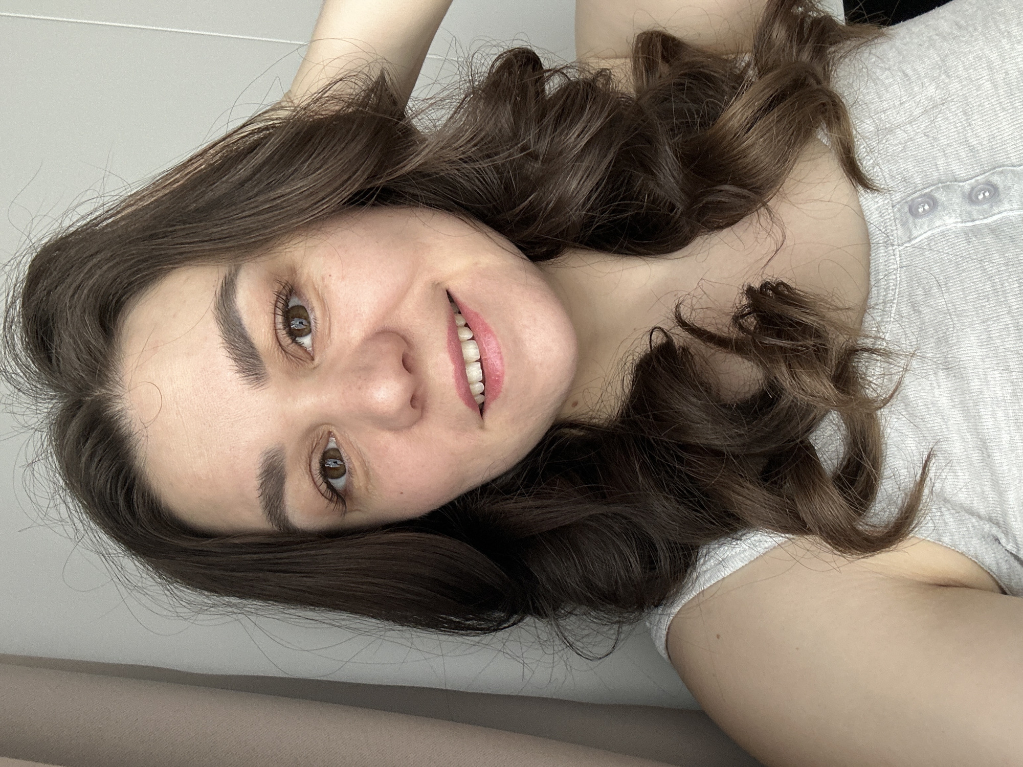 This is a close-up, tilted selfie of a young woman, likely taken indoors in Chaikovsky, Russia. The woman is smiling genuinely, looking directly at the camera with warm, brown eyes. Her lips are slightly parted, revealing white teeth, and she is wearing light pink lipstick. Her eyebrows are dark and well-defined.
She has long, voluminous, dark brown hair with noticeable curls and waves, which frames her face and spreads around her. She appears to be lying down or leaning back, with one arm subtly visible at the top of the frame, possibly resting behind her head. She is wearing a simple, light grey or off-white t-shirt with two small, light-colored buttons visible near the neckline.
The background is minimalist, consisting of a plain, light-colored wall in the upper portion and a darker, soft brownish surface in the lower portion, suggesting a cozy, personal setting like a bedroom or living room. The lighting is soft and even, highlighting her features and creating a relaxed atmosphere. There is no visible text in the image. Her expression conveys happiness and comfort.