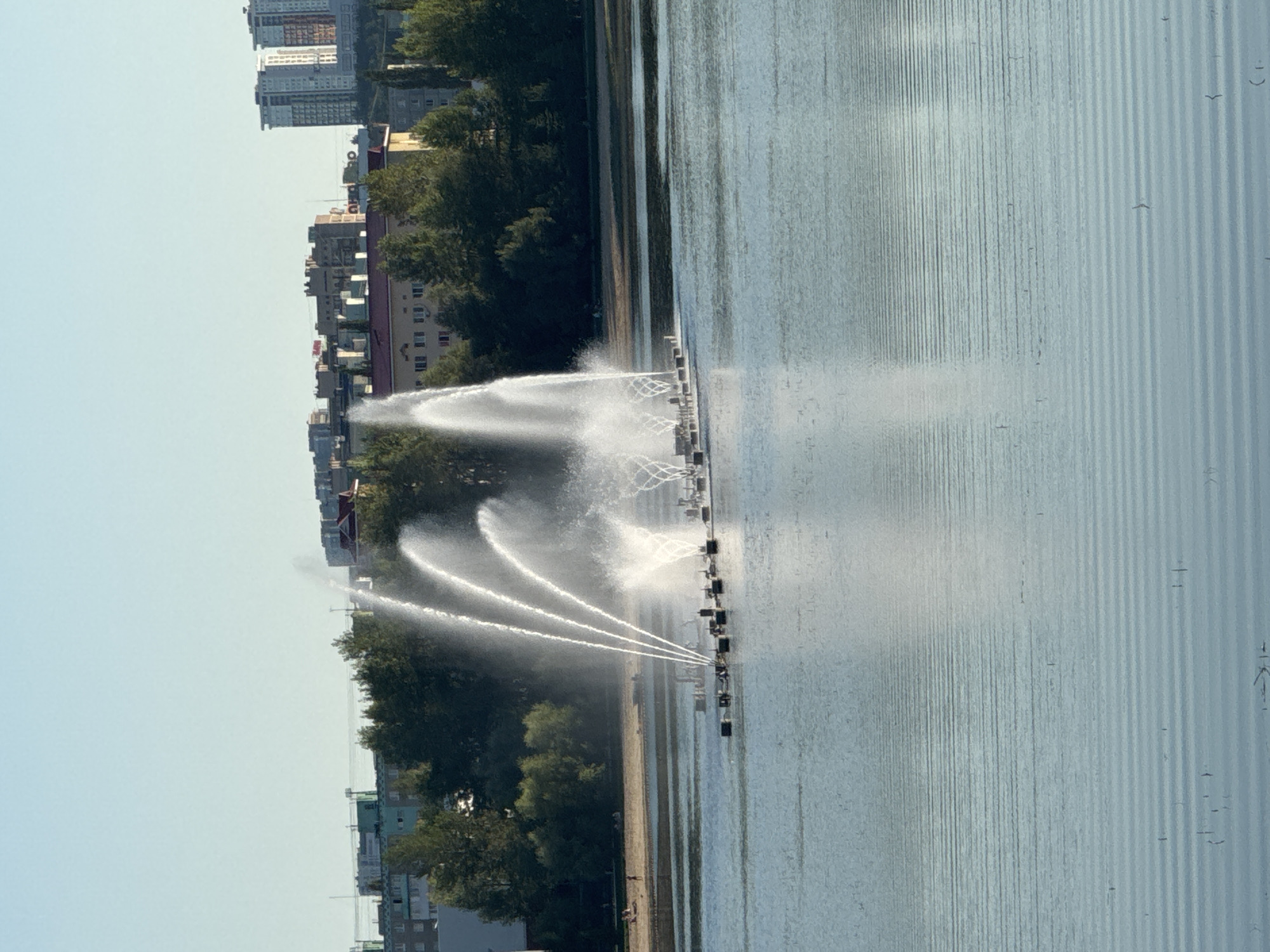 The image depicts a wide-angle view of a lake with an active fountain in the center. The fountain is comprised of multiple jets of water spraying upwards, creating a dynamic visual display. In the background, a treeline separates the water from a city skyline characterized by apartment buildings of varying heights and architectural styles, with some under construction. The sky is clear and a pale blue. The water surface exhibits a subtle ripple effect, reflecting the light. A sandy area, possibly a beach or park, is visible along the edge of the lake, with a few individuals casually present. This scene is identifiable as the "Yuzhny" (Southern) district of Ufa, Russia, specifically the lake and its surrounding embankment which is a popular recreational spot.