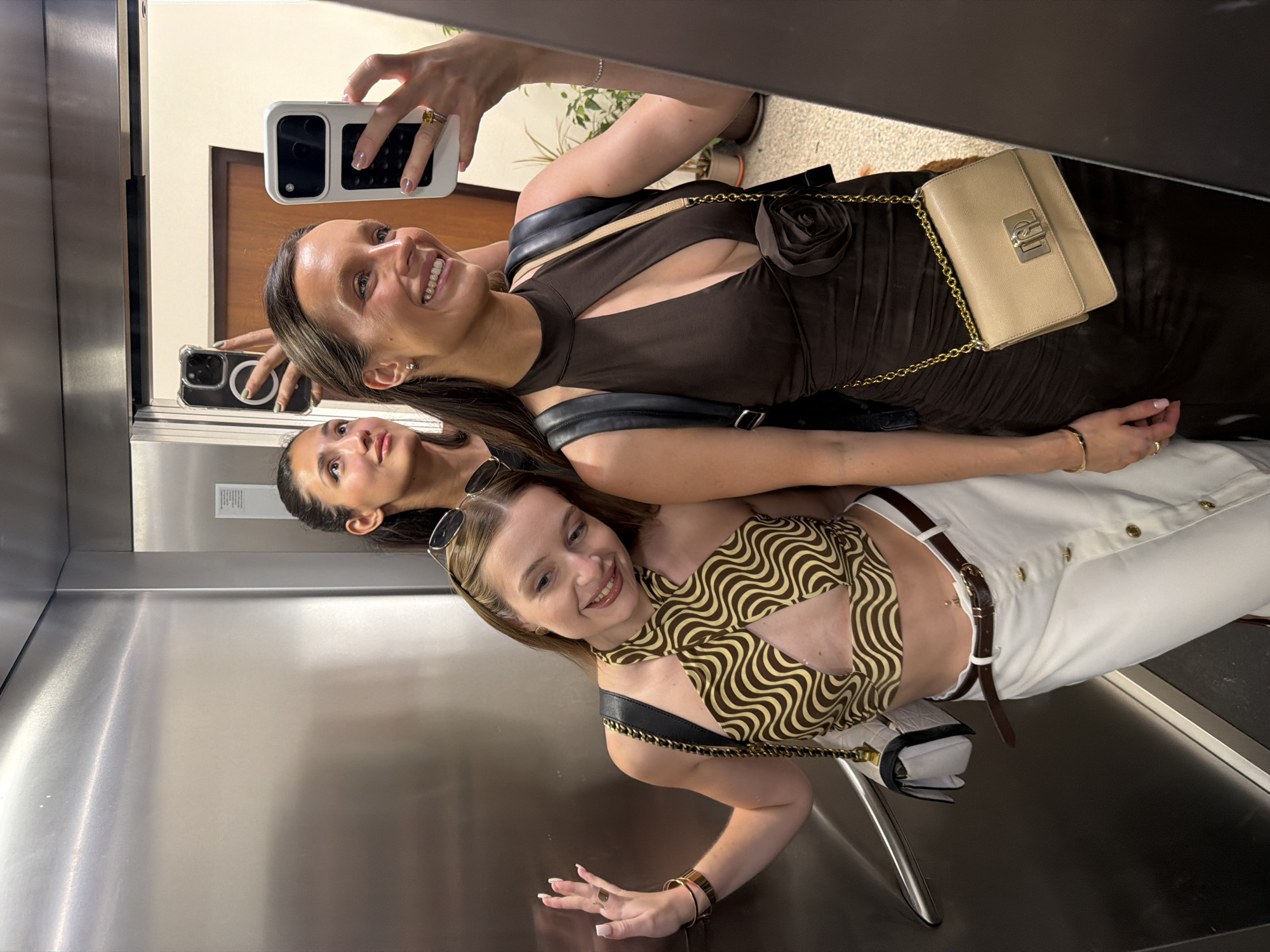 The image displays three young adult females inside an elevator, specifically capturing a mirror selfie, in Budva, Montenegro.
The individual on the far left, a fair-skinned female with light brown hair and sunglasses resting on her head, smiles directly at the camera. She wears an olive green and brown wavy-patterned halter neck crop top with a central chest cut-out, a white midi skirt with a brown button-front closure, and a dark brown belt. A navel piercing is visible. Her left wrist features a gold-toned bracelet, and a black and white quilted shoulder bag with a gold chain strap is carried on her left side.
Adjacent to her, a second female with dark hair is partially visible, primarily in the mirror's reflection. She looks upward with a slight smile, holding a dark-colored smartphone with a clear MagSafe-compatible case overhead.
The third individual, positioned on the far right and taking the photograph, presents a frontal view with dark hair and a wide smile. She wears a dark brown, sleeveless, high-neck dress with a keyhole cut-out at the chest, ruched detailing, and a dark brown fabric rose embellishment on the torso. Her accessories include small silver stud earrings, a thin gold bracelet on her right wrist, a yellow-stone ring on her right ring finger, and a beige shoulder bag with a gold chain and clasp, worn over a dark backpack. She holds a white smartphone with a dark grip case.
The elevator interior features a brushed metallic left wall and a large mirrored right wall. The mirror reflects the three females and a background beyond the elevator doors, which includes a light brown door frame, a cream-colored wall, and several potted green plants on a light-colored tiled floor.