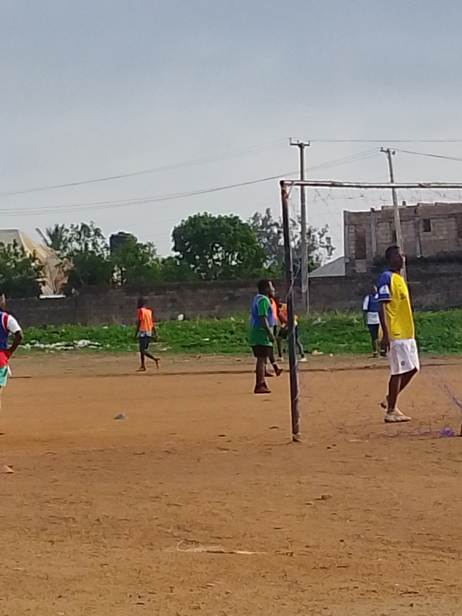 The image shows a soccer field in Bukuru, Nigeria. There are several young men on the field, some wearing athletic attire and likely playing or training. One man is wearing a yellow and blue jersey with white shorts, standing near a makeshift goal post. Other men are wearing vests of various colors including orange, green, and red. The field is dirt, with sparse grass patches in the background, along with some shrubs and trees. Behind them, there is a partial view of some buildings, one of which is under construction. Overhead power lines are visible. The sky is overcast.