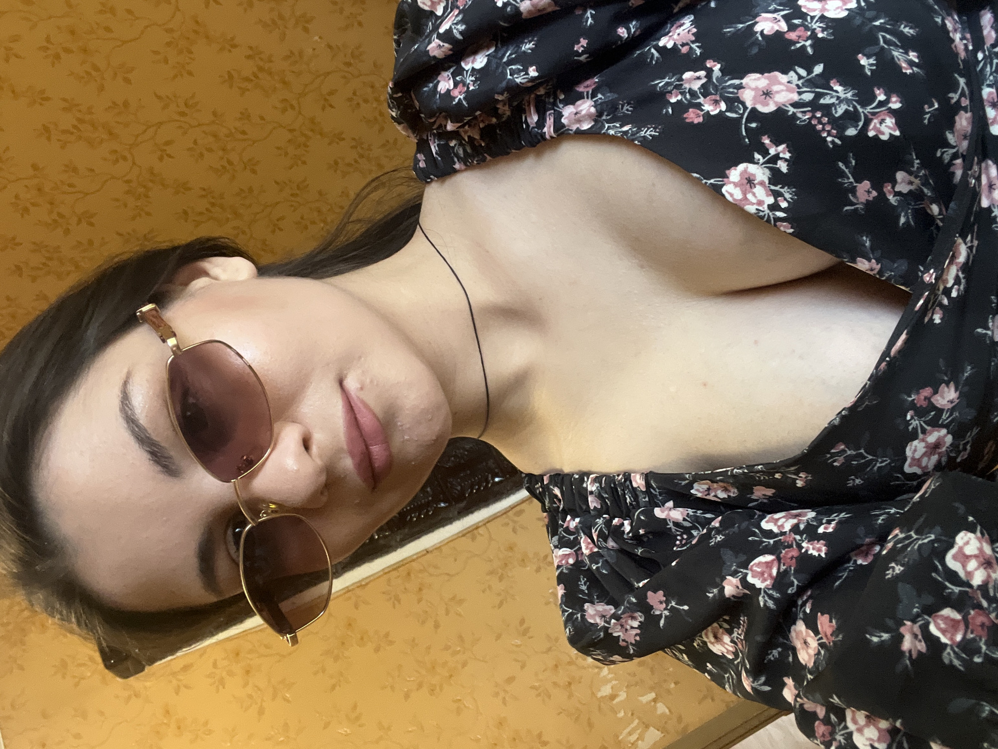 A young woman with dark hair, wearing sunglasses and a floral dress, poses for a selfie against a textured yellow wall. The woman is looking at the camera, with her head slightly tilted. The floral dress is black with pink and white flowers. The background wall has a repeating pattern of branches and leaves.
