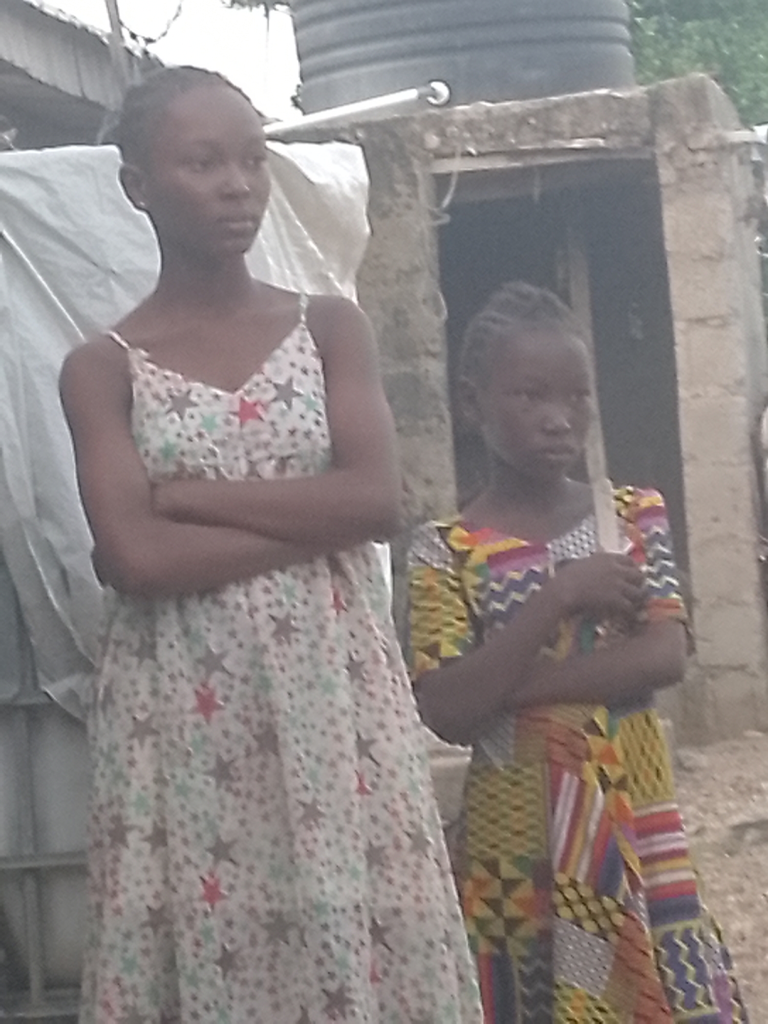 Two girls stand with arms crossed in front of a building, in the city of Unknown, Unknown. The girl on the left wears a white sundress patterned with stars, while the other is clad in a colorful, patterned dress. In the background, a water tank is visible, along with part of a corrugated metal structure, and the stone or concrete building they are standing near.