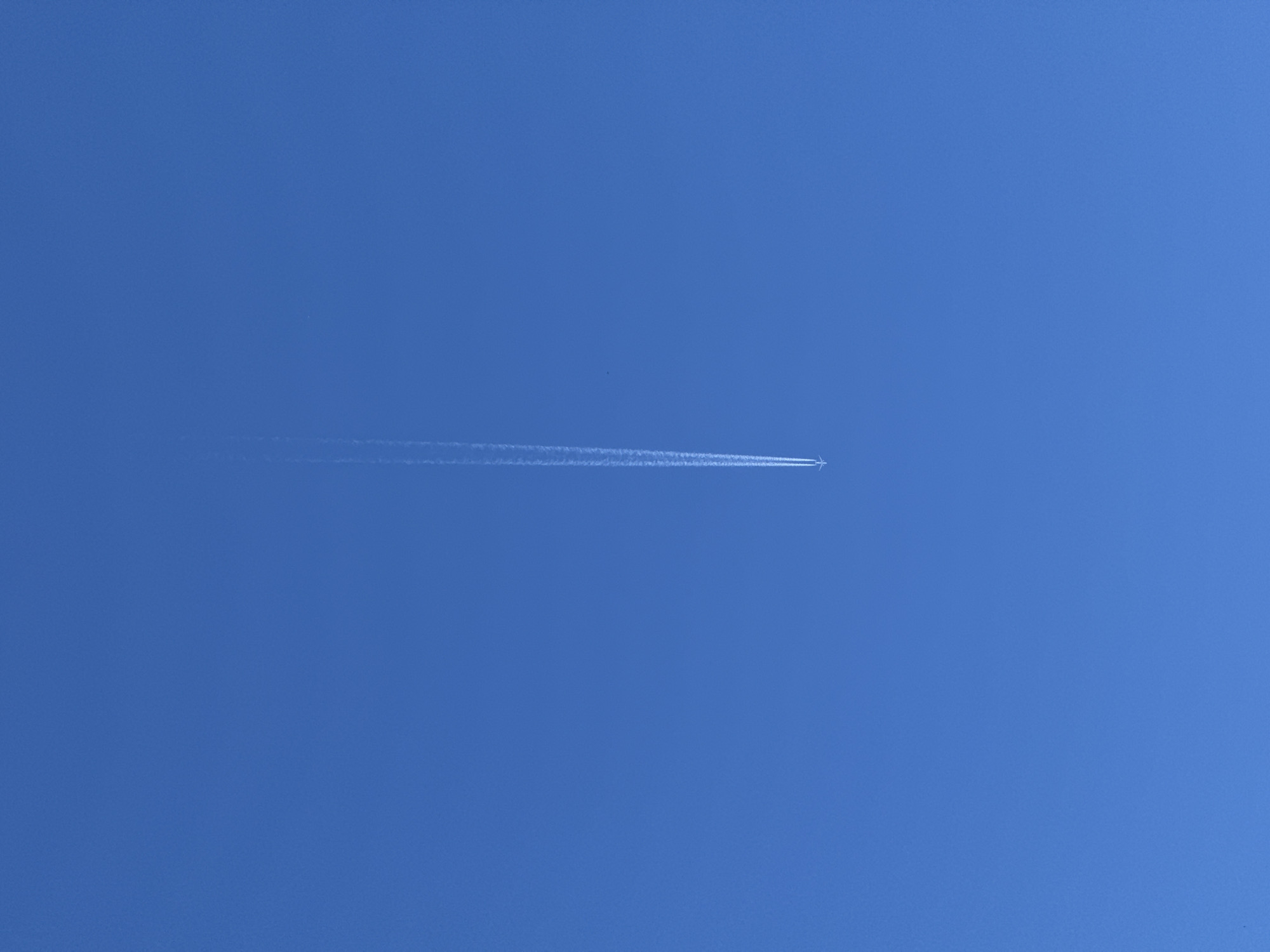 An airplane leaves a white contrail as it flies across a clear, bright blue sky. The airplane is small and appears at the bottom center of the image. The contrail stretches upwards from the plane, tapering off as it ascends. The image is devoid of any landscape or city elements, focusing solely on the aircraft's passage through the atmosphere. It is not possible to determine the location in the city of Slavyani, Bulgaria from this image.