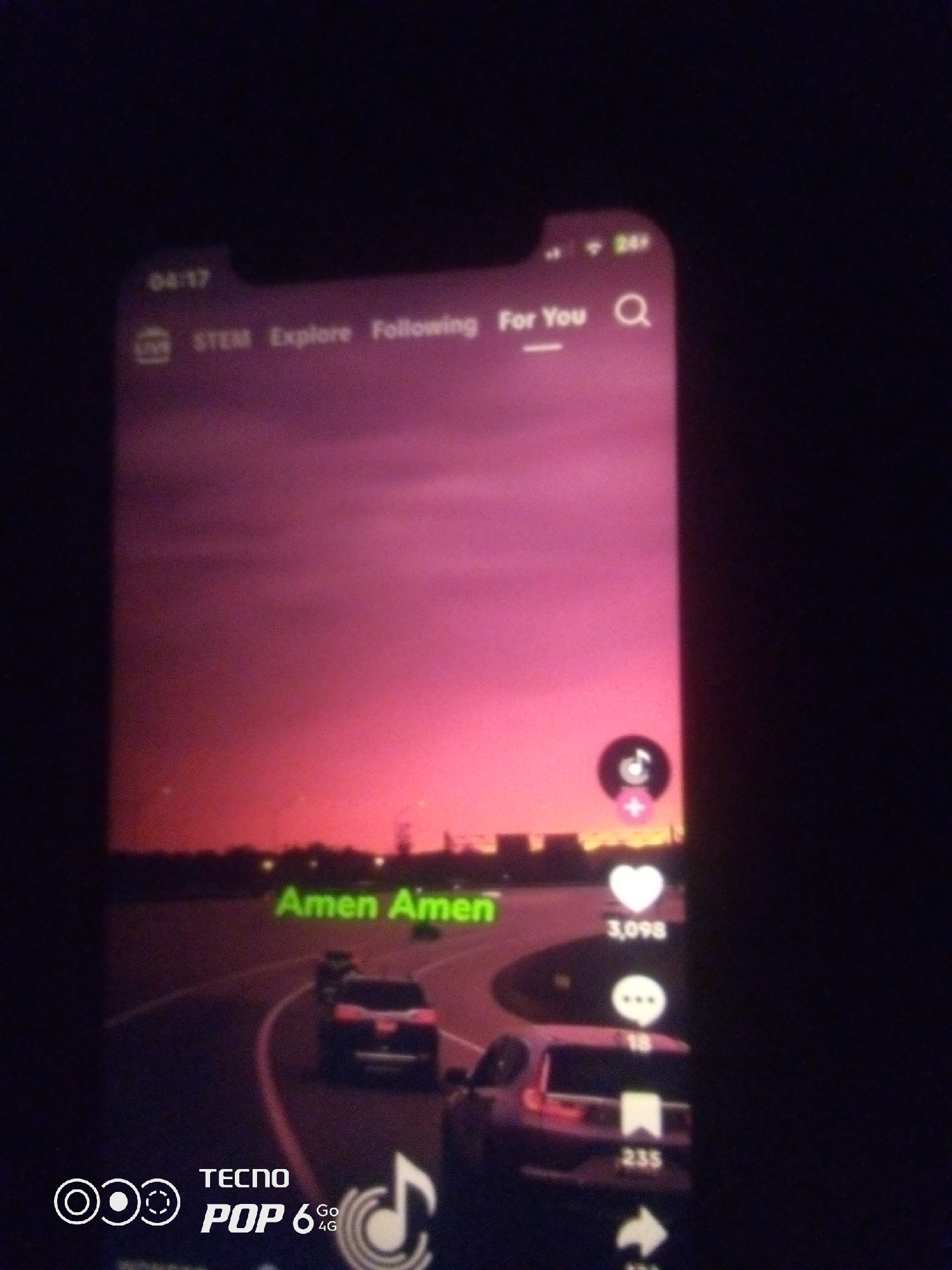 The image shows a screenshot of a mobile phone displaying a social media video feed, likely TikTok, given the interface elements. The video features a sunset with a vibrant pink and purple sky, silhouetted city lights in the distance, and a highway with several cars driving. Text overlayed on the video reads "Amen Amen." On the right side of the screen are standard social media interaction icons: a heart with the number 3,098, a chat bubble with the number 18, and a bookmark icon with the number 235. At the bottom of the phone screen, there is branding for "TECNO POP 6 4G". The phone itself appears to be on a dark surface, and the lighting suggests it's nighttime or low-light conditions. There is no visible indication of the specific location in Howayi, Nigeria.