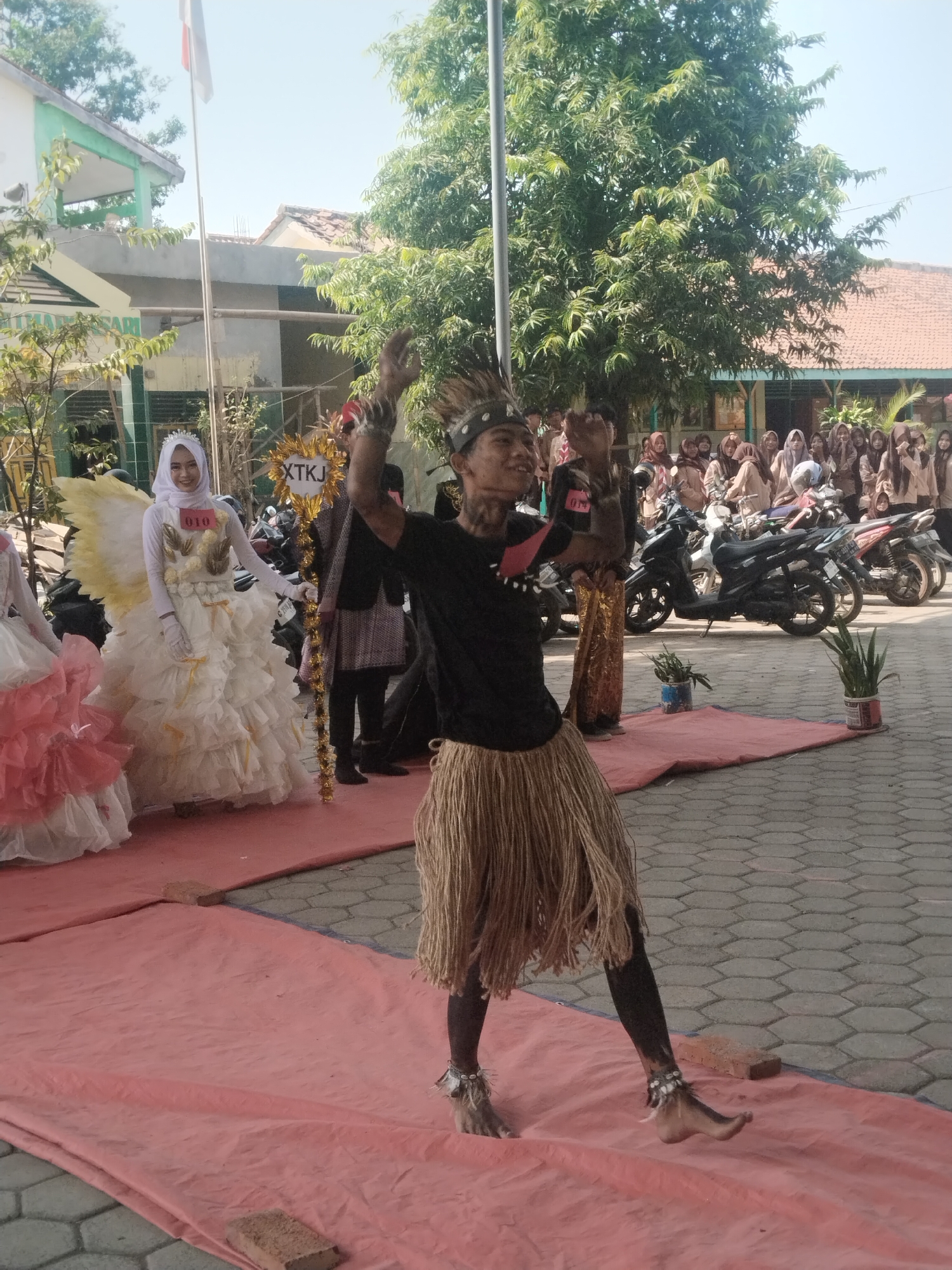 In Pakulaut, Indonesia, on a paved ground covered with a pink tarp, a young male is performing a dance. He is wearing a headdress made of animal fur, a black t-shirt, and a skirt made of dried grass. His legs are painted black, and he wears anklets with shells. He has his arms raised, his hands open, and his body is in a dynamic pose, suggesting movement. In the background, a white-clad person with large yellow wings and a frilly dress is standing. Behind them, a group of young people in uniform are observing the performance. Motorbikes are parked in the background. The scene is set outdoors, with trees and buildings suggesting a school or community gathering place. The sky is bright blue, indicating a sunny day.
