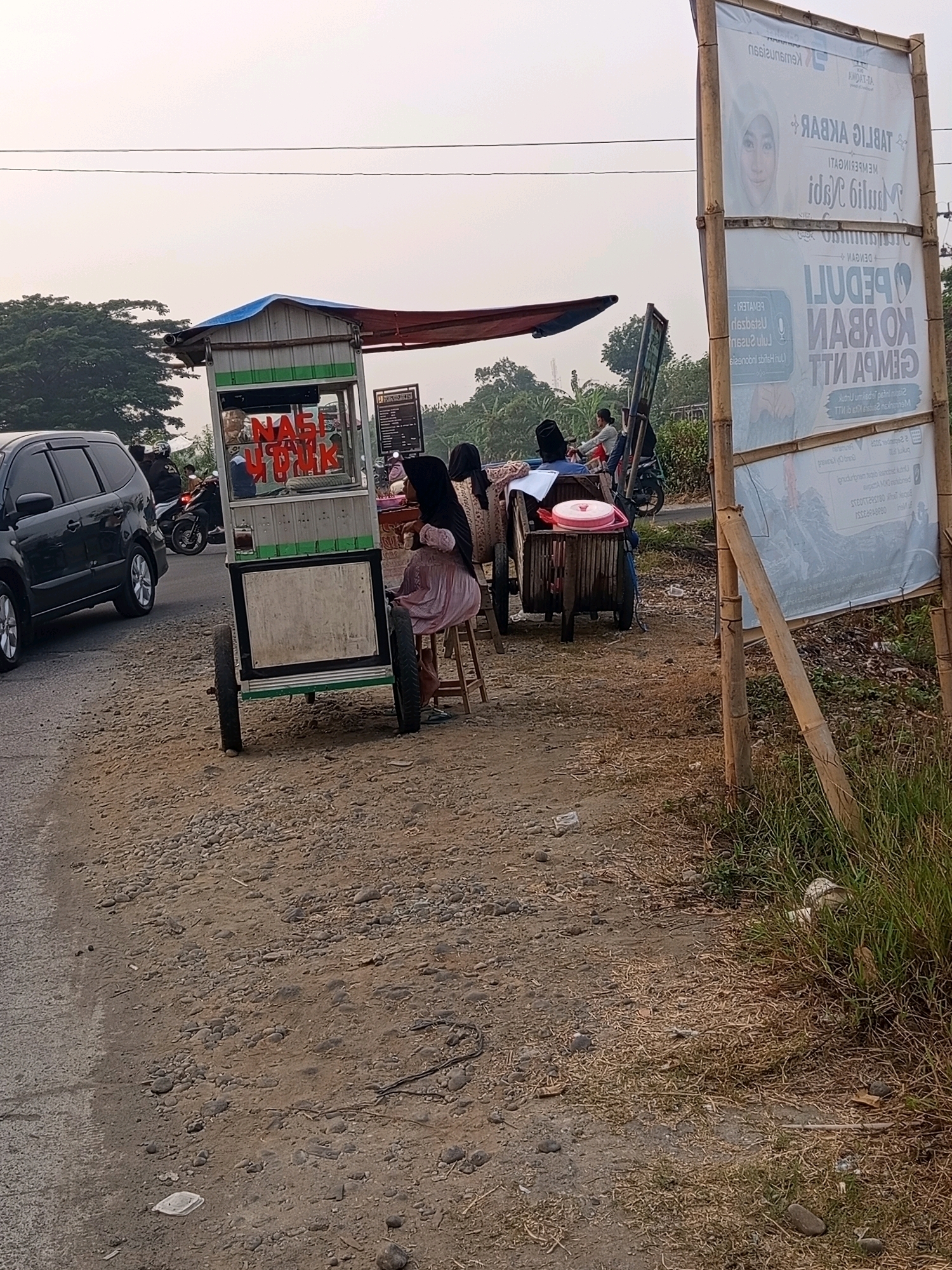 The image depicts a roadside scene in Karawang, Indonesia, likely on a street with moderate traffic and pedestrian activity. In the foreground, a food cart with red and white signage, displaying the words "Nasi Uduk," is parked on a gravelly roadside. Two individuals, one possibly a vendor and the other a customer, are positioned near the cart. The customer, wearing a headscarf and a light-colored dress, is seated on a stool, facing away from the camera. Further down the road, a black car is visible, and several motorcycles are parked or in motion, suggesting a dynamic environment. A large banner, displaying text and an image of a woman's face, is propped up by bamboo poles on the right side of the frame. The background consists of greenery and a hazy sky, characteristic of an outdoor setting in Indonesia. The overall scene suggests a typical day in Karawang, with local commerce and daily life unfolding along the roadside.