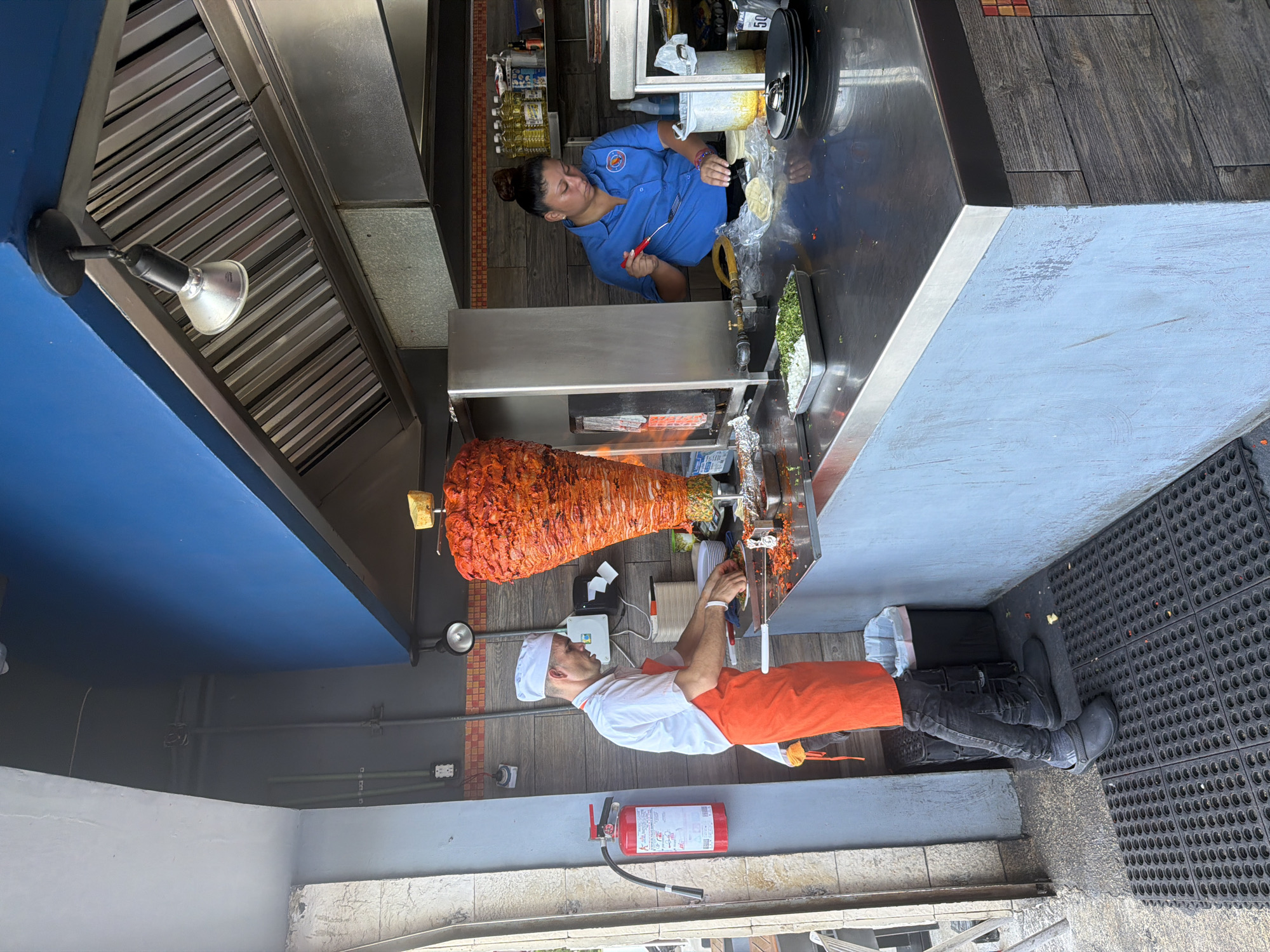 In Cancun, Mexico, a busy taqueria is depicted. Two individuals are actively preparing food behind a stainless steel counter. On the left, a man wearing a white chef's hat, white shirt, and an orange apron is carving from a vertical rotisserie filled with marinated meat, likely al pastor. He is focused on his task, with plates and ingredients laid out before him. To the right, a woman in a blue shirt is working with tortillas and what appears to be a container of salsa or liquid, using a red utensil. The background reveals a commercial kitchen setup with shelves holding bottles, a ventilation hood, and a fire extinguisher mounted on the wall. The taqueria features a vibrant blue wall and a rustic, tiled base for the counter, suggesting an authentic, local dining establishment.