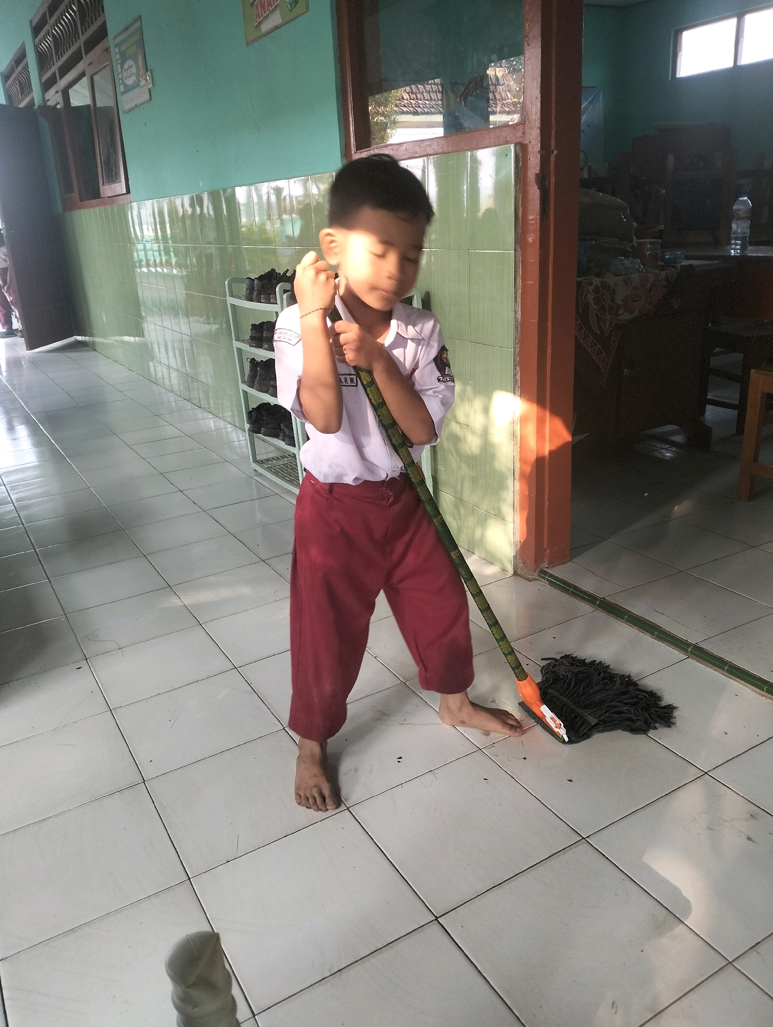 A young boy, wearing a white school shirt with a red collar and red pants, is mopping a tiled floor in what appears to be a school hallway. He is holding a mop with a dark fabric head and a green and brown patterned handle, which is positioned on the floor. The boy's right hand is on the mop handle, and his left hand is near his face. He is barefoot. The hallway walls are painted a light green, and there are windows on the left side of the image. A shoe rack with several pairs of shoes is visible on the wall behind the boy. The floor tiles are white with dark grout lines. Sunlight streams in from the right, creating shadows on the floor. The scene is located in Blabakan, Indonesia, likely within an educational facility.