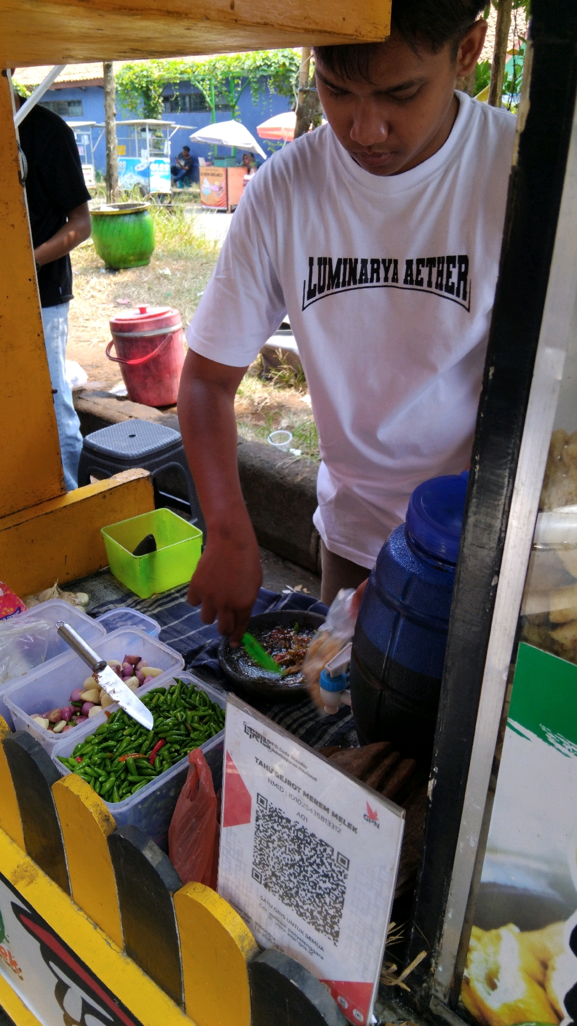 In Proyonangg, Indonesia, a young male wearing a white t-shirt with the text "LUMINARYA AETHER" printed on it is seen standing behind a food stall. The stall has yellow and black painted wooden fencing. In front of him, there are various containers filled with ingredients. One container holds chopped shallots and garlic, another contains numerous small green chilies, some with red tips. To the right, a mortar and pestle is being used by the young man, suggesting he is preparing food. A large blue water dispenser is also visible. A QR code is displayed on a stand near the front of the stall. In the background, other food stalls and people can be seen under umbrellas, indicating a bustling outdoor market environment.