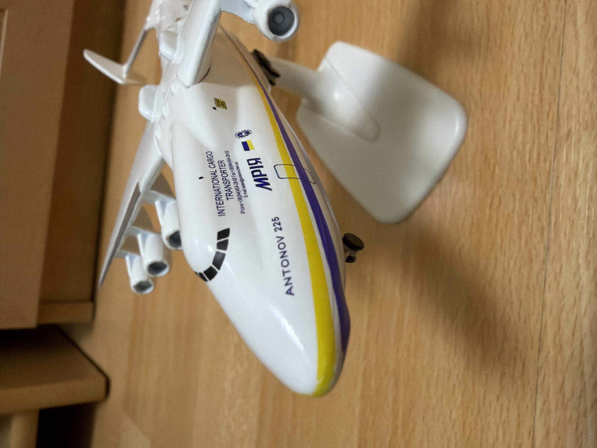 A model of an Antonov An-225 Mriya aircraft is displayed on a wooden surface. The model is white with blue and yellow stripes along its fuselage and wings. Text on the fuselage reads "INTERNATIONAL CARGO TRANSPORTER," "ANTONOV 225," and "МРІЯ." The background shows a blurred view of wooden furniture. There are no people or actions depicted. The image does not provide information about the city of Kyiv, Ukraine, beyond the aircraft's association with Antonov, a Ukrainian manufacturer.