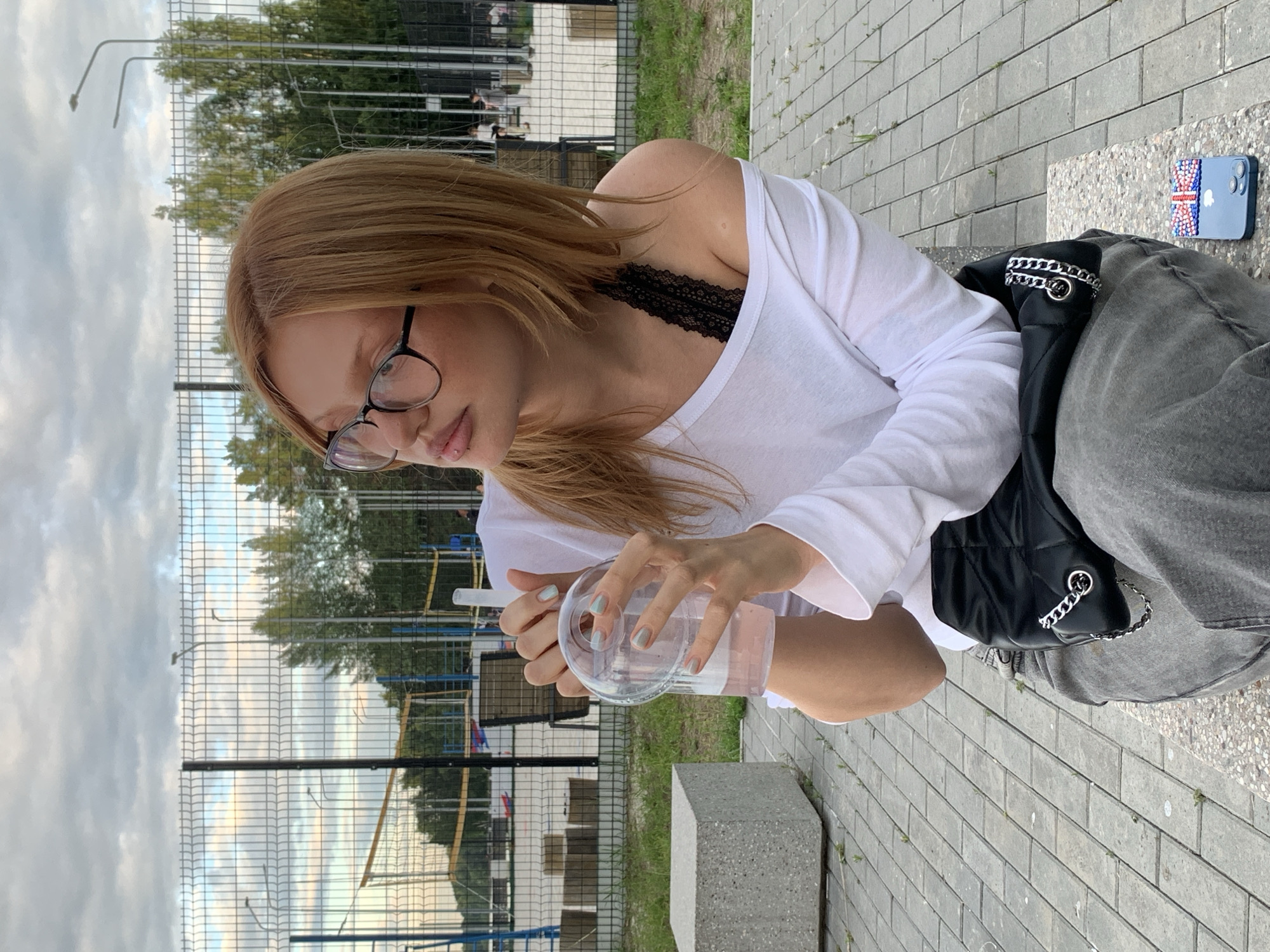 A young woman with blonde hair and glasses is sitting outdoors. She is wearing a white off-the-shoulder shirt and a black lace bra. She is holding a clear plastic cup with a straw and appears to be drinking from it. Her nails are painted a light blue-green color. A black quilted bag with a silver chain strap is resting next to her, and she is wearing grey jeans. On a bench to her right, a light blue phone case decorated with the Union Jack flag is visible.
The background shows a chain-link fence, trees, and a cloudy sky, suggesting an overcast day. The ground is paved with gray bricks. The overall scene appears to be a park or outdoor recreational area, possibly a sports court given the fence. The woman's gaze is directed downwards, and her expression is neutral. The image is taken in portrait orientation. No visible text is present in the image.