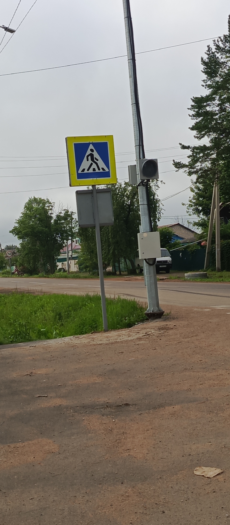 The image shows a pedestrian crossing sign on a pole, a traffic signal, and a speed limit sign in the city of Pogranichny, Russia. The pedestrian crossing sign is yellow and blue with a white silhouette of a person walking. The traffic signal is black and mounted on the same pole as the pedestrian crossing sign. A speed limit sign with the number "50" is visible in the background to the left. The scene appears to be on a cloudy day, with lush green grass and trees surrounding the road. A white van is parked on the side of the road in the background.