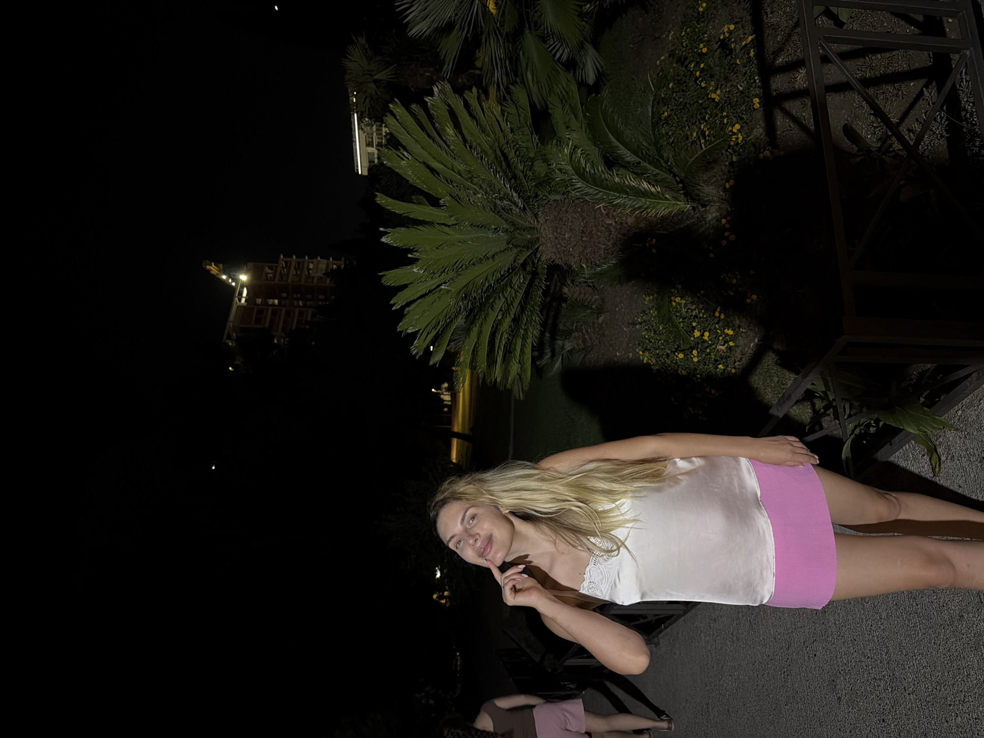 A blonde woman is posing for a photo at night in Budva, Montenegro. She is wearing a white satin camisole with lace trim and a pink mini skirt. Her left hand is raised to her chin, and she is smiling at the camera. In the background, a construction site with a crane is visible, along with palm trees and other greenery. The scene is dimly lit, suggesting it is late evening or nighttime.