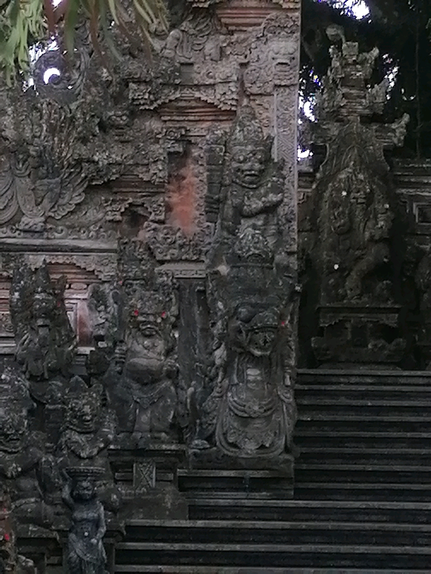 The image shows a stone relief carving of a temple entrance, likely in Ubud District, Indonesia. The carving features a series of deities or guardians, each with intricate headdresses and stern expressions. Two large, imposing figures flank the entrance, their bodies appearing muscular and adorned with elaborate ornamentation. Smaller figures are carved below them, some carrying objects on their heads. Steps lead up to the temple, also carved from stone. Lush green foliage frames the top of the image, suggesting an outdoor setting. The stone appears weathered, indicating age and exposure to the elements.