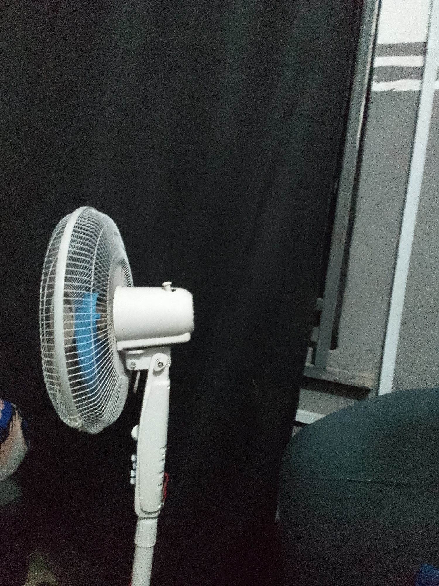 A white pedestal fan is shown in a dark room. The fan has a white cage and a blue interior. The fan is on a white stand, which is partially obscured by a dark object in the foreground. To the right of the fan, a gray wall with white stripes is visible. The lighting in the room is dim.