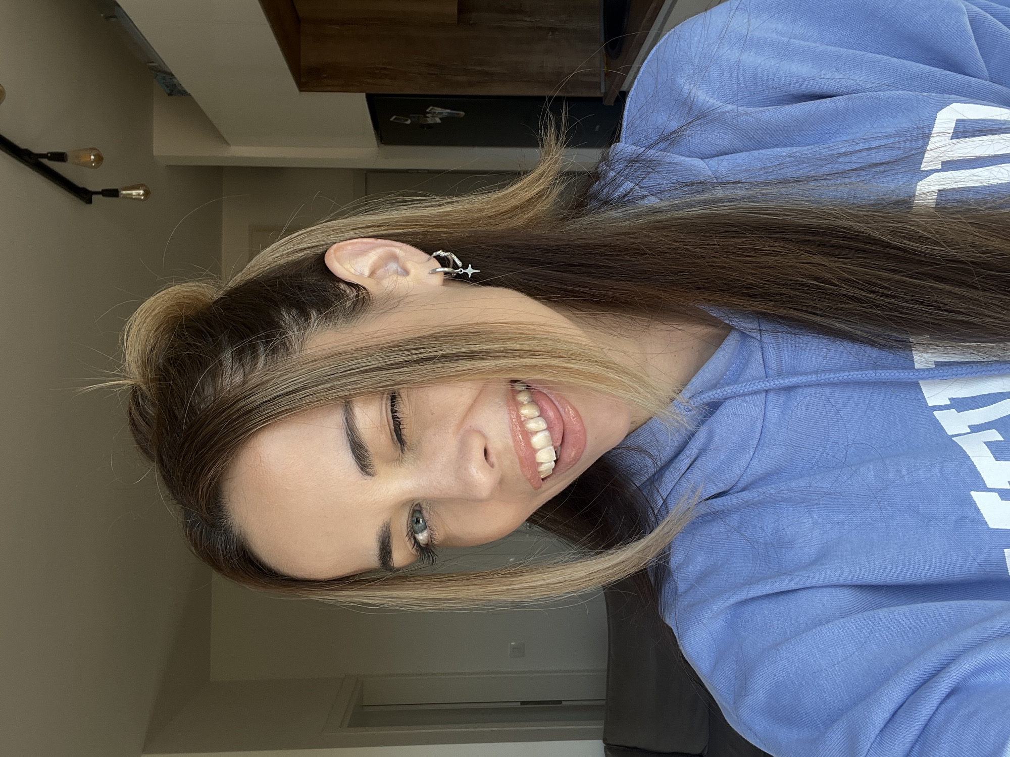 A young woman with long, straight, light brown hair, highlighted with blonde tones, poses playfully for a photo in an indoor setting. She is positioned diagonally, tilting her head to her left, with a wide, cheerful smile that reveals her upper teeth and a glimpse of her tongue. Her left eye is open, while her right eye is partially closed in a wink.
She is wearing a light blue hooded sweatshirt (hoodie). Large white letters are visible on the front of the hoodie, including a distinct uppercase "E" and partial subsequent letters. In her visible left ear, she wears a silver-colored ear cuff adorned with a small star dangle and another curved design. Her facial features include well-defined eyebrows and lips with a glossy finish.
The background depicts a casual indoor environment. To the left, a light-colored wall features a black, industrial-style light fixture with two exposed filament bulbs. Above, a dark wooden beam or shelf is visible against a lighter ceiling. The lighting is soft and even. While the specific location cues for Bar, Montenegro, are not discernible, the general indoor setting is consistent with such a location. The overall mood conveyed is happy and playful.