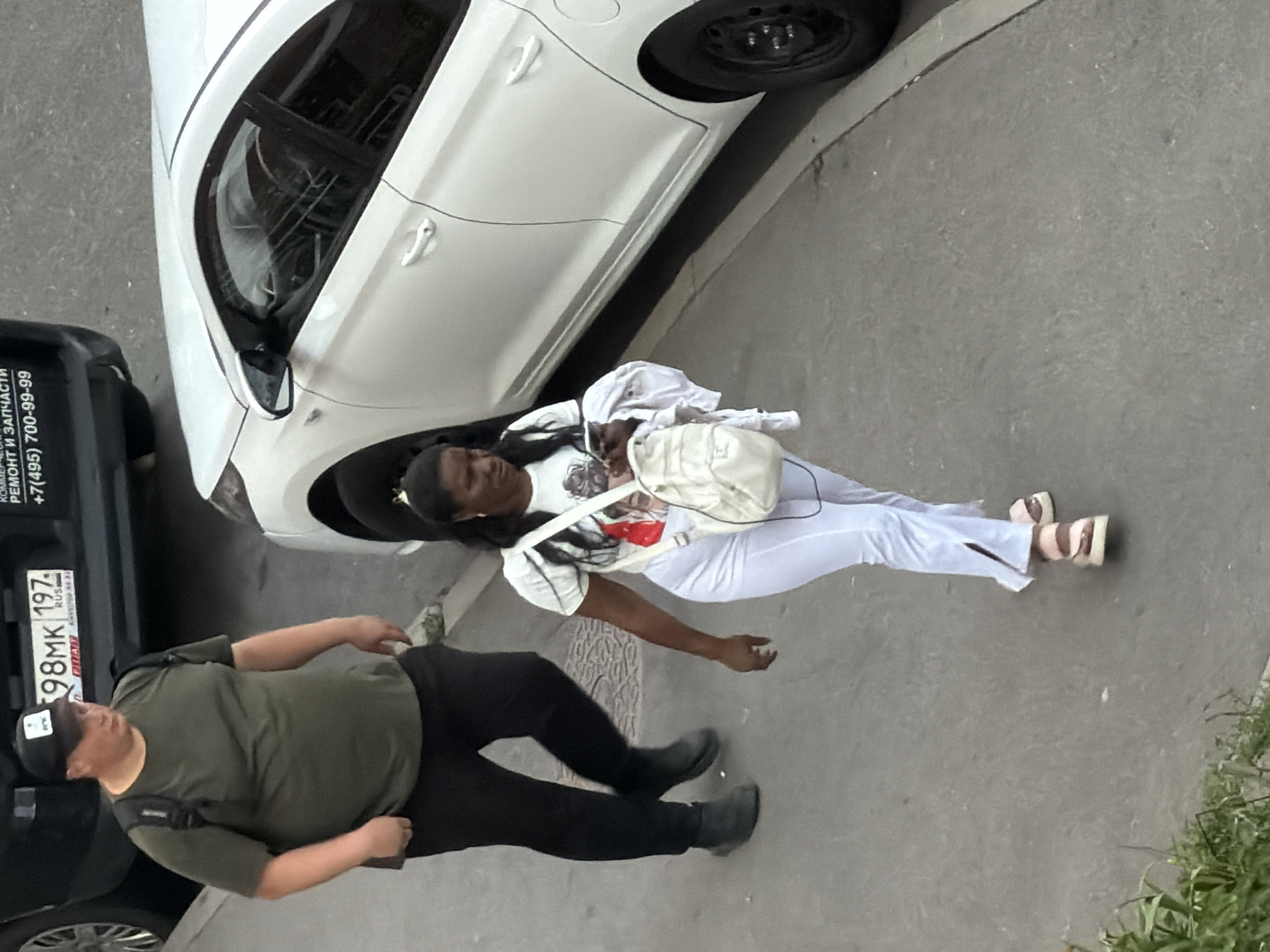 A medium shot from a high angle shows two individuals walking on a paved surface in an urban environment. On the left, a heavyset male wearing a dark green t-shirt, black pants, and a black baseball cap with a logo walks away from the camera. He has a black backpack on. To his right and slightly ahead, a woman with dark hair and earrings, wearing a white t-shirt with a graphic print and white pants, walks towards the camera. She is carrying a white backpack on her left shoulder and a white garment in her right hand, and is looking down at her phone. A white car is parked to the right of the woman, with its side facing the viewer. Part of a dark vehicle is visible on the far left. The ground is a textured grey pavement with some green vegetation visible in the lower left corner. The image appears to be taken during daylight.