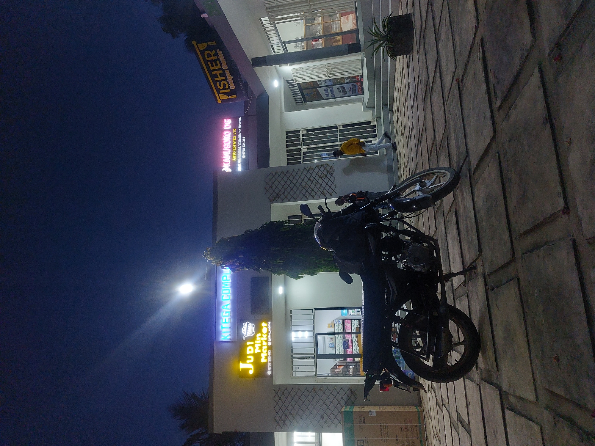 The image depicts a nighttime scene in Dar es-Salaam, Tanzania, in front of a commercial building with multiple businesses. A parked motorcycle is in the foreground, oriented diagonally.
The businesses visible include "Judi Mini Market" and "Kitega Complex," indicated by illuminated signs. A sign for "Fisher Unique Center" is also visible. Other signs mention "Kamusoko DG" and "Auto Estates Ltd", listing services related to land/property.
A man is visible near the entrance of one of the businesses, wearing a yellow top. The environment suggests a paved area in front of the building. Some packaged goods can be seen near the motorcycle, possibly awaiting transport. The lighting is a mix of natural darkness and artificial illumination from the business signs and building lights.