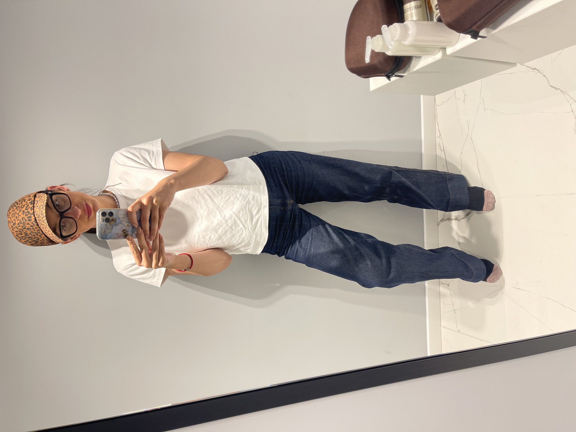 A woman takes a selfie in a bathroom mirror. She is wearing a white t-shirt, blue jeans, a leopard print headscarf, and glasses. She is holding a cell phone in her hands. The bathroom has gray walls, a white marble countertop, and a black-framed mirror. There is a brown bag and some bottles on the countertop. The image does not provide any information to identify the location as being in Sofia, Bulgaria.