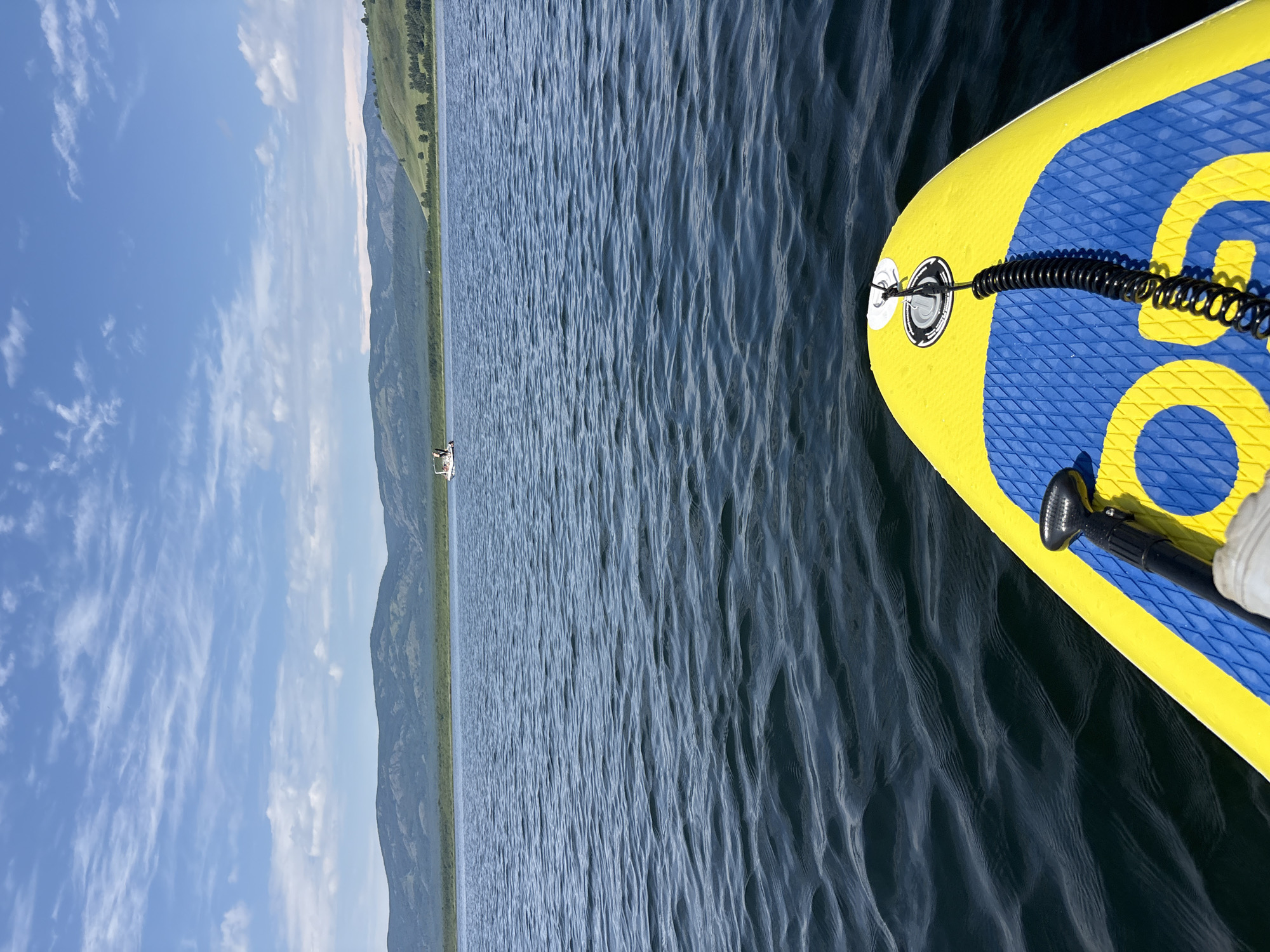 The image shows a first-person perspective from a yellow and blue paddleboard on a calm lake. The paddleboard has a coiled leash attached to it. In the distance, a small white boat with two people is visible on the water. Beyond the lake, there are green hills and mountains under a blue sky with scattered white clouds. The scene suggests an outdoor recreational activity on a body of water. The location of Кусимовского рудника, Russia is not discernible from the image itself.