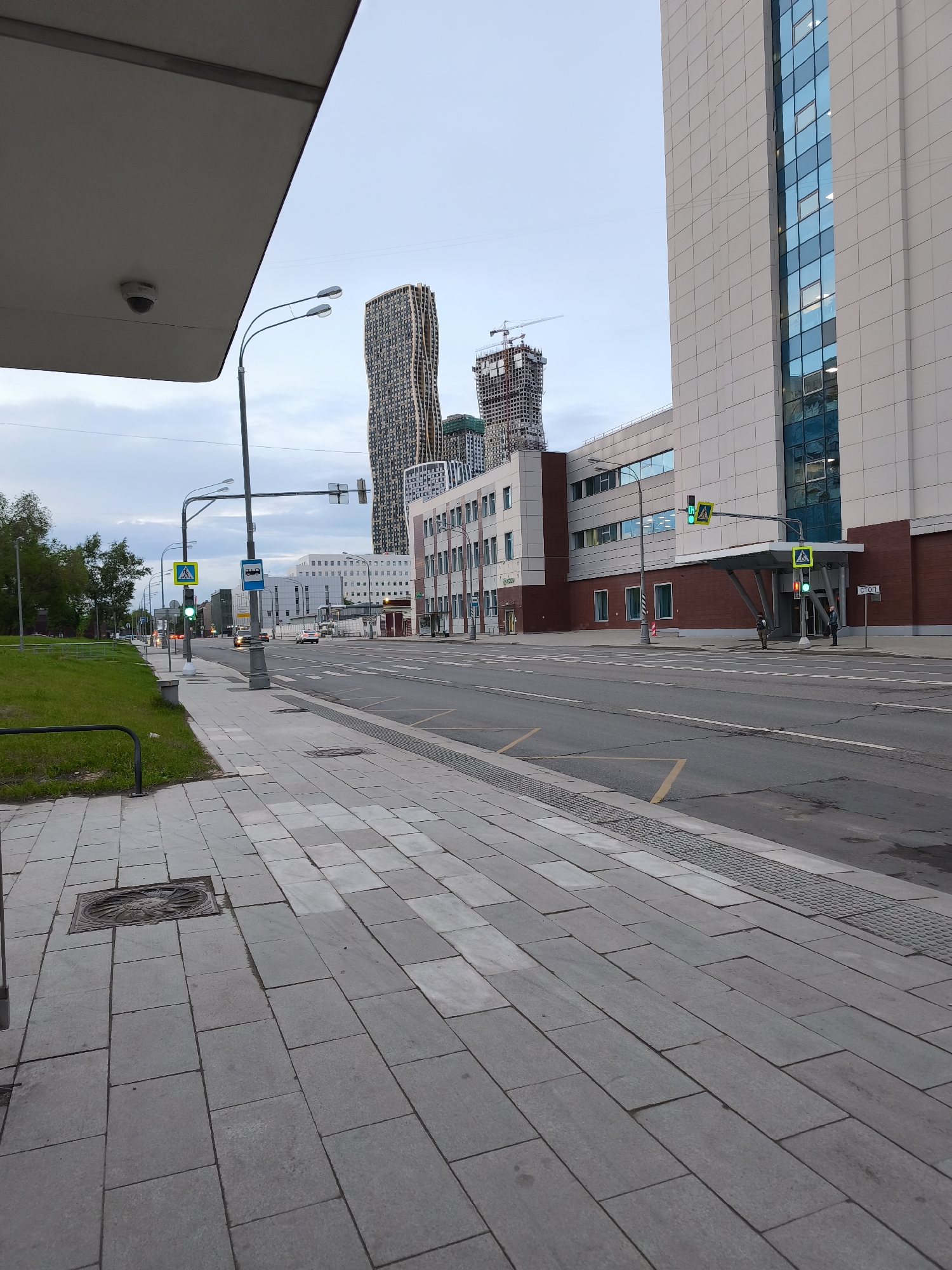 The image shows a street scene in Moscow, Russia. A wide, mostly empty street divides the scene. On the right is a modern building, clad in light-colored panels with blue-tinted windows. A second building under construction is to the right of that. Overhead a canopy, a street light, and a traffic light with a green pedestrian signal are present. On the left is a sidewalk paved with gray rectangular stones. Green grass is along the left edge. Two people stand near a building. A bus stop is visible further down the street, and several cars drive in the distance.
