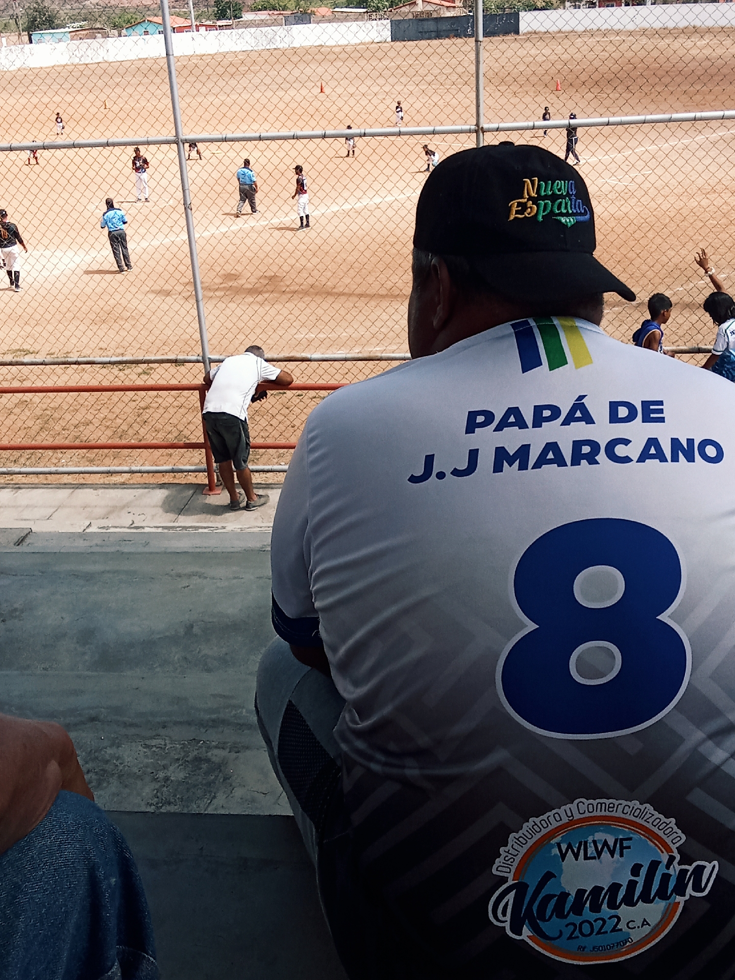 This is a medium shot taken at a baseball field, seemingly in Juan Griego, Venezuela. In the foreground, a man wearing a "Nueva Esparta" baseball cap and a custom shirt that reads "Papa de J.J Marcano" with the number 8 on the back is sitting. The shirt also has a logo at the bottom that reads "WLWF Kamilin 2022 C.A." Another man is leaning against the chain-link fence on the opposite side. In the background, a baseball game is in progress on a dusty field. Several players are visible in different positions, suggesting ongoing gameplay. The background also includes what appears to be a residential or urban area, indicating that the field is located within a populated region.