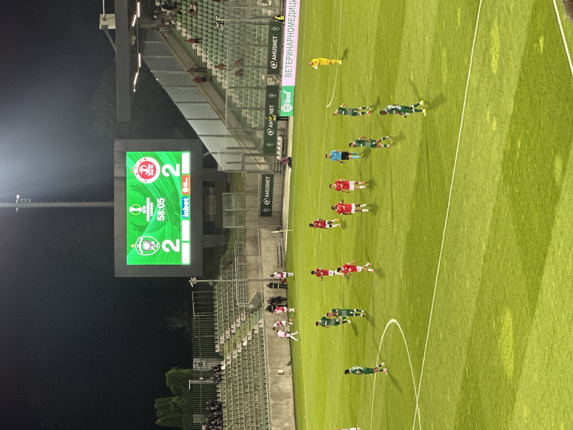 The image shows a night-time view of a football match at the Huvepharma Arena in Razgrad, Bulgaria. A large scoreboard in the background displays the score as 2-2 and the time as 58:05, indicating a Conference League match. Players in red and green kits are visible on the brightly lit green football pitch, engaged in play. Spectators are seated in the stands, which are illuminated by stadium lights. The overall scene captures a moment of athletic competition in a modern sports venue.