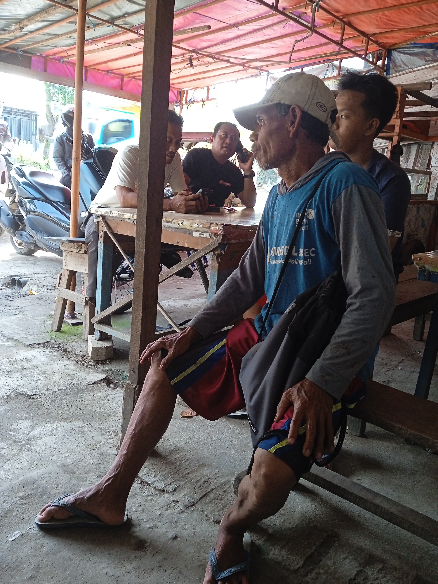The image captures a candid scene in Sekopek Wetan, Indonesia. Four men are present under a corrugated metal roof structure. One man is seated on a wooden bench in the foreground, wearing a cap, long-sleeved blue shirt, red and blue shorts, and flip-flops. He has a black bag slung over his shoulder. Three men are seated at a wooden table; one is on a cell phone. A blue motorcycle is parked nearby. The ground is concrete, and the overall environment appears to be a roadside eatery or gathering place.